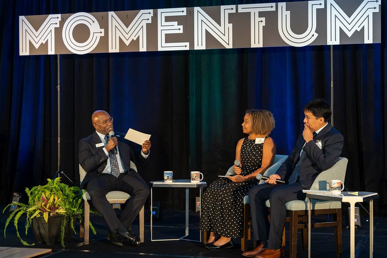 Three people seated on a stage beneath a large 'MOMENTUM' banner during a panel discussion.