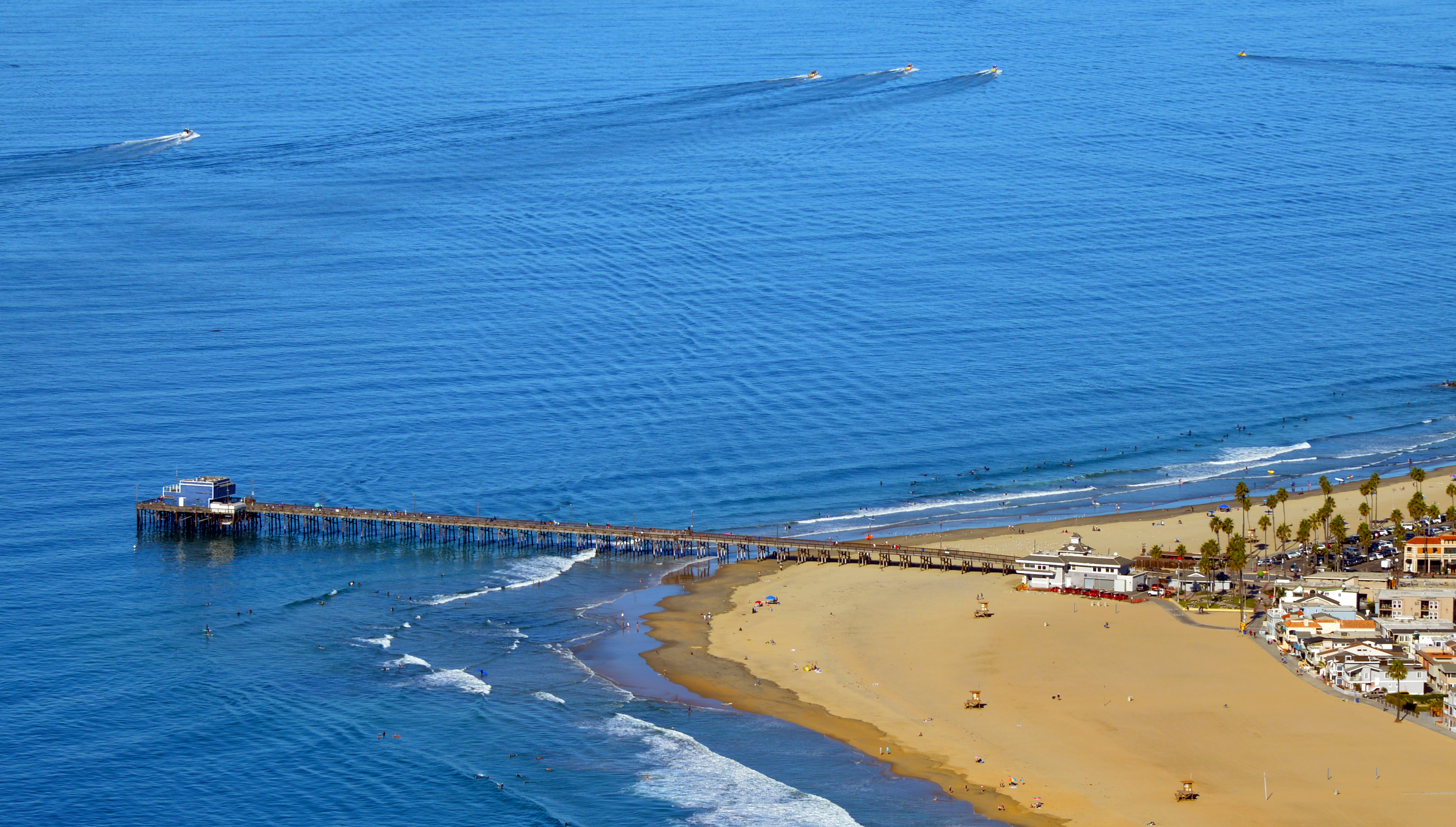 Aerial view of a pier extending into the ocean with a sandy beach and coastal buildings below.
