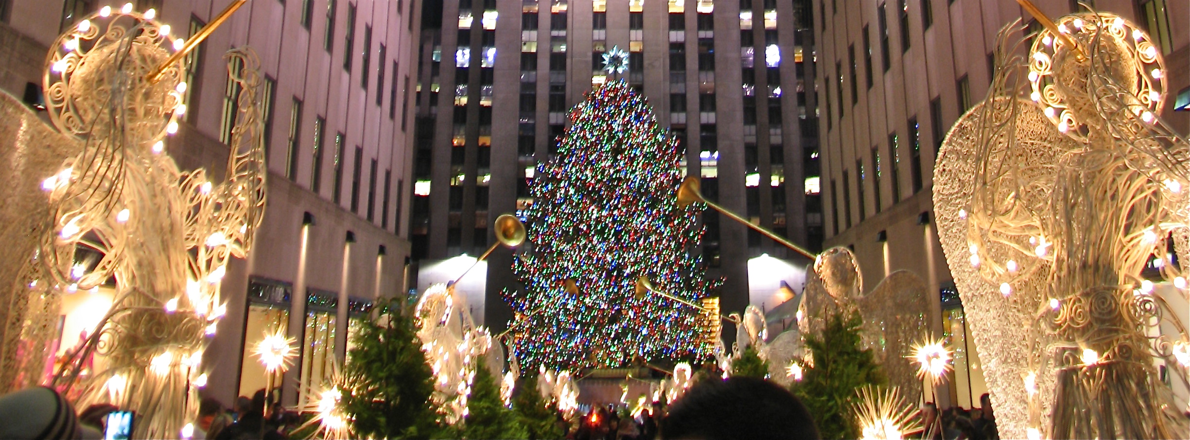 Illuminated Christmas tree flanked by glowing angel sculptures at night, with crowds and a skyscraper in the background.