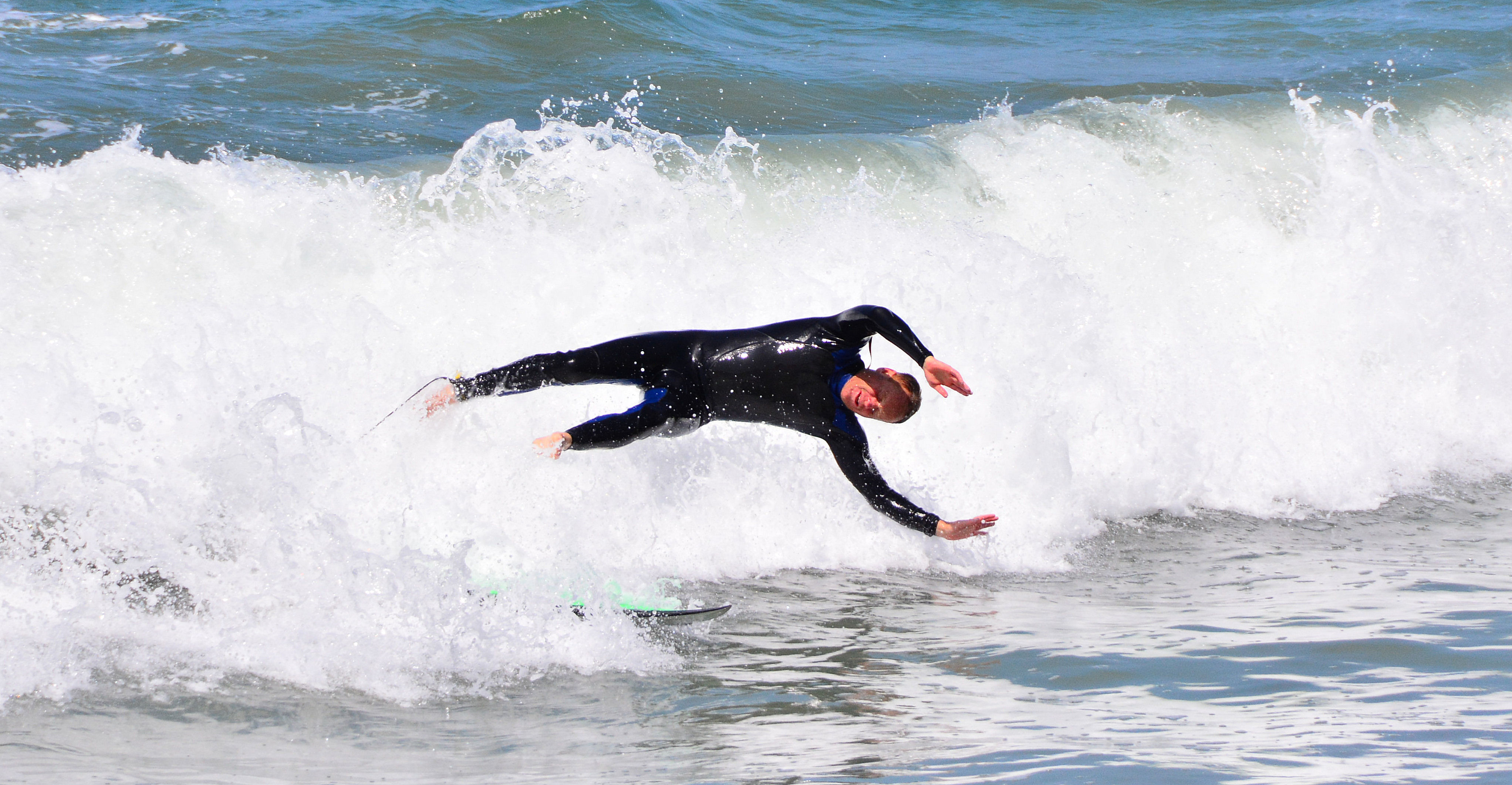 A surfer in a black wetsuit tumbles off a surfboard on a crashing wave.
