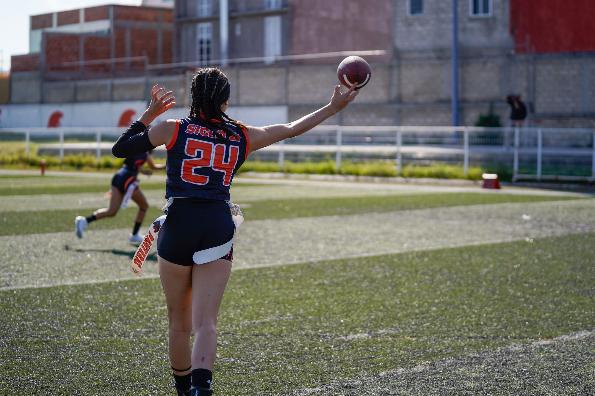 Two female athletes playing flag football on grass field in sunny weather.