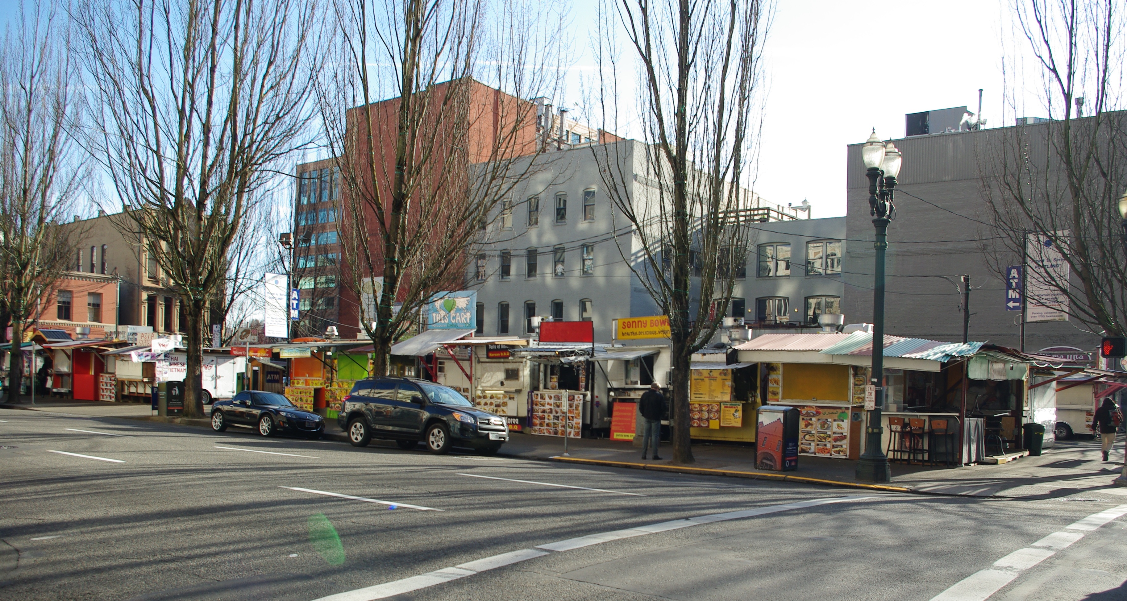 Multiple food carts in an urban pod with bare trees and parked vehicles along a downtown Portland street.
