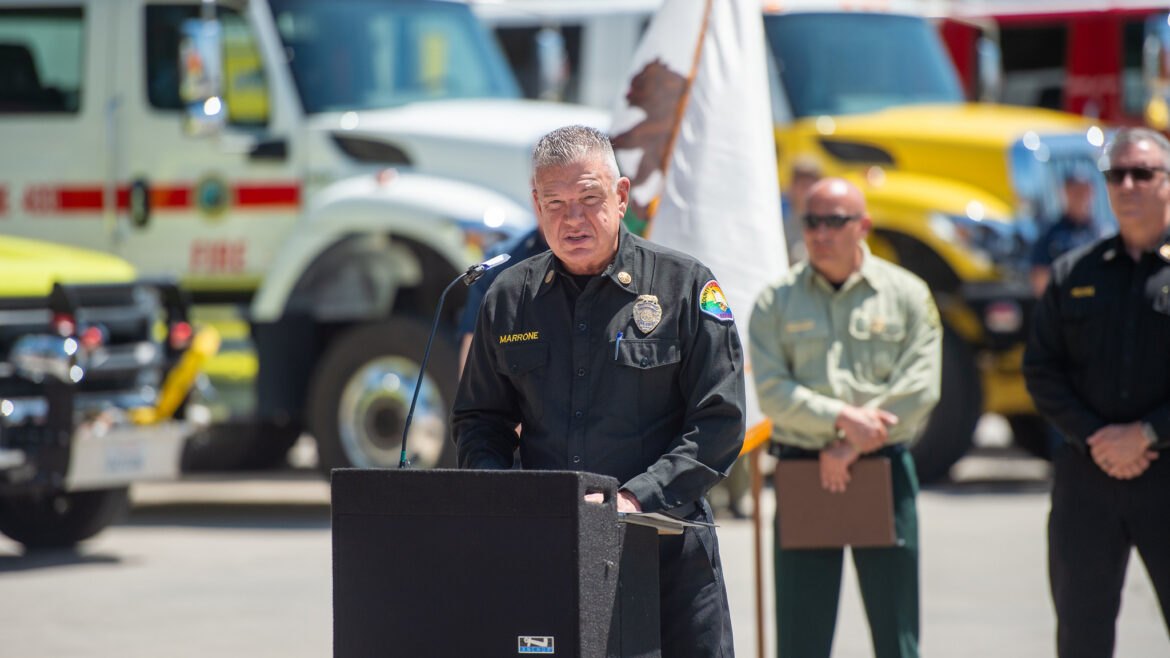 Fire official in dark uniform speaking at outdoor podium with fire trucks behind.