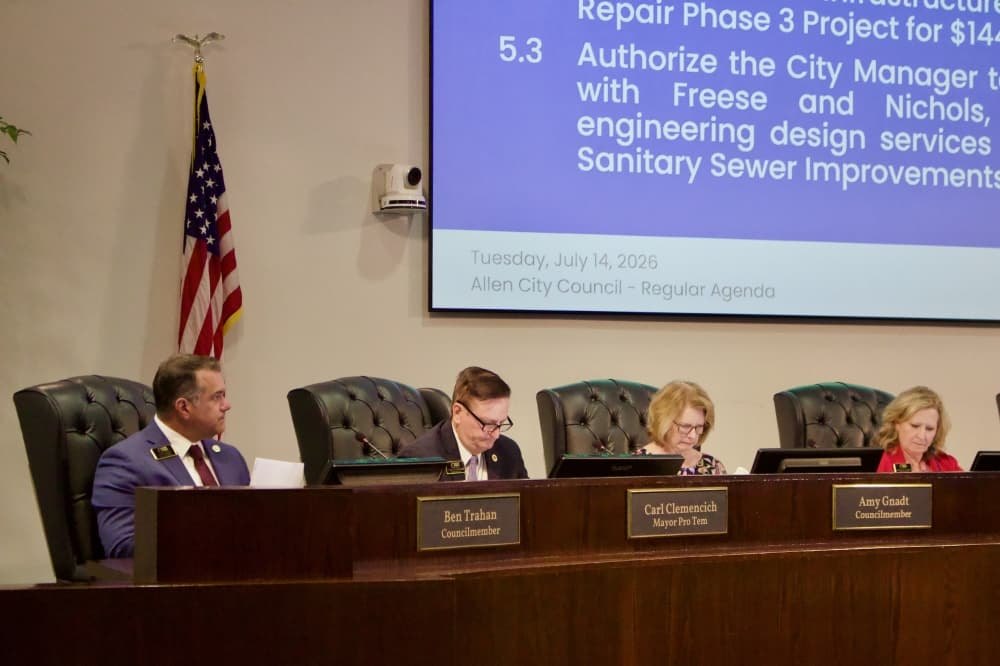 Four council members seated at a curved dais with a projected agenda screen behind them.