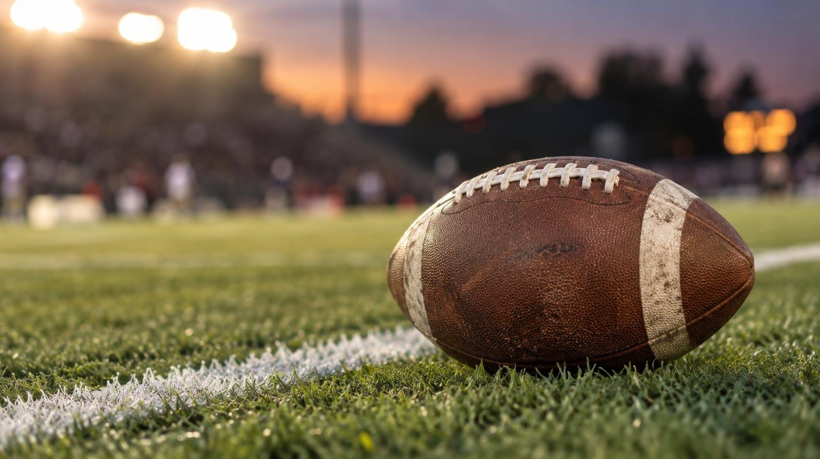 An American football resting on green grass beside a white yard line under stadium lights.