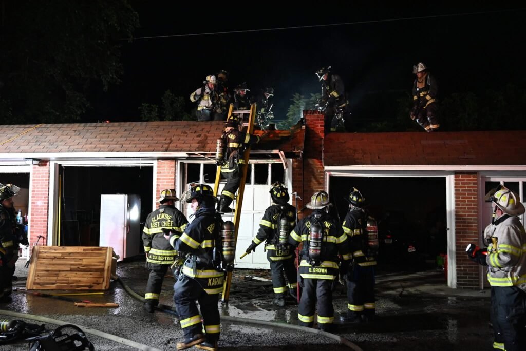 Firefighters in turnout gear on the roof and ground level of a brick garage row at night, smoke rising above.