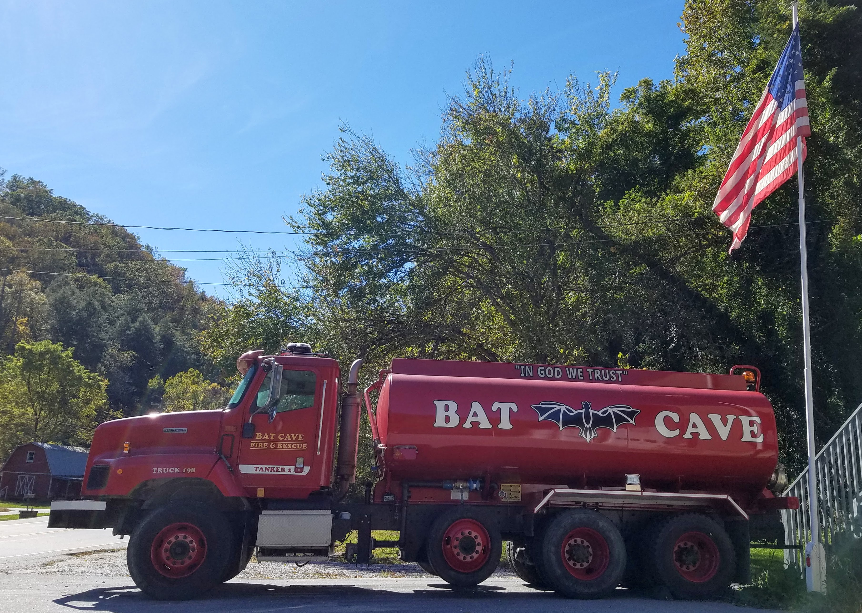 A red tanker fire truck parked outside a volunteer fire station with a flagpole nearby.