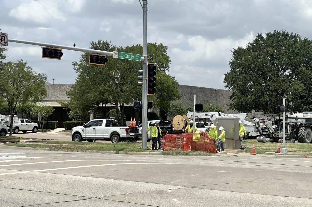 Construction workers in high-visibility vests work near a traffic signal at a street intersection.