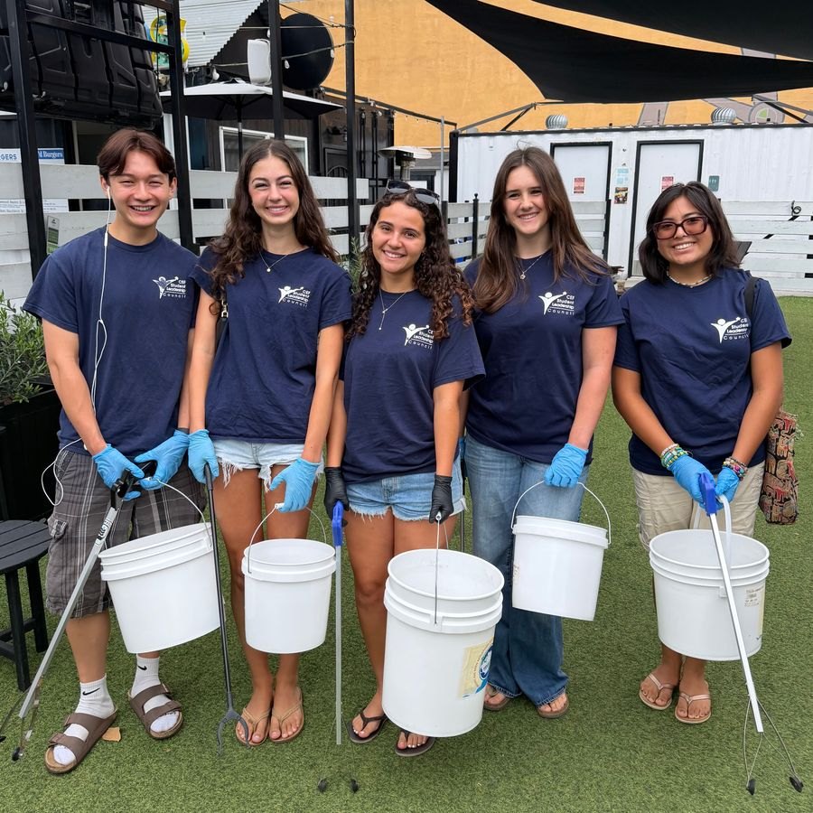 Five young volunteers in navy shirts and blue gloves hold white buckets and litter-picking tools outdoors.
