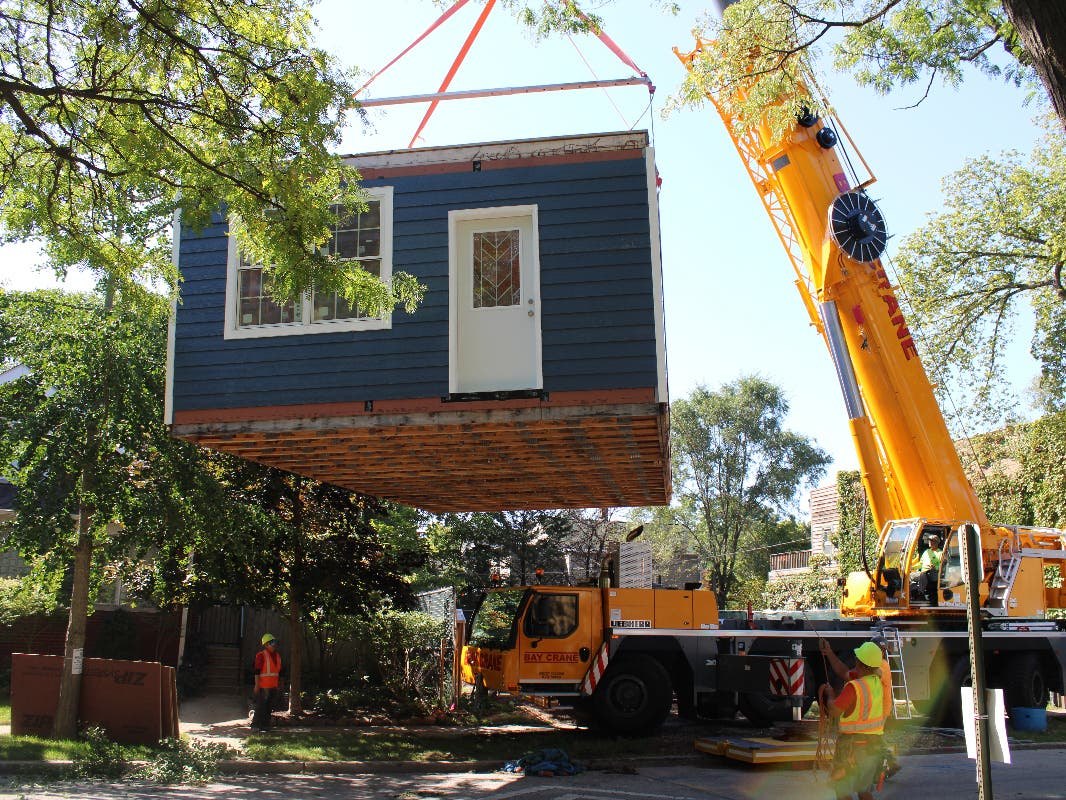 A small two-story house with blue siding is lifted by a large yellow crane.