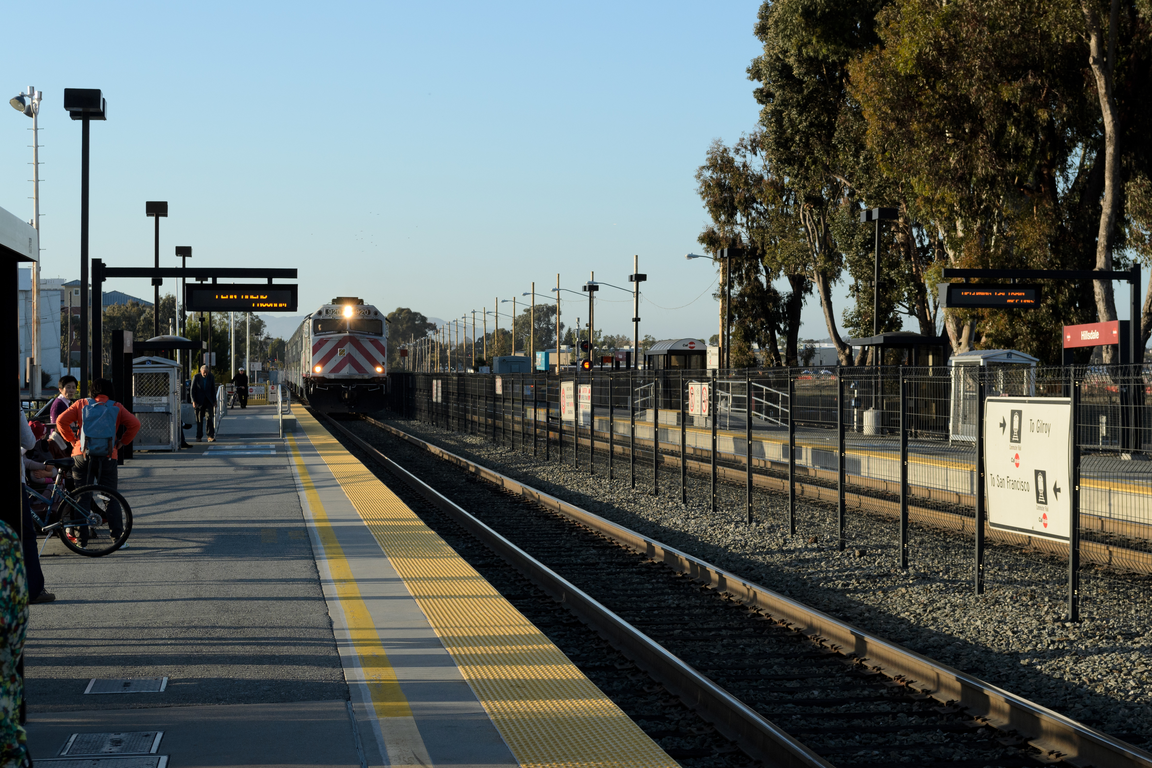 A Caltrain train approaches a station platform with passengers and bicycles waiting.