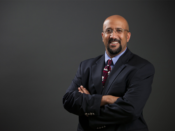 Bald man with glasses and beard in dark suit, arms crossed, smiling against gray background.
