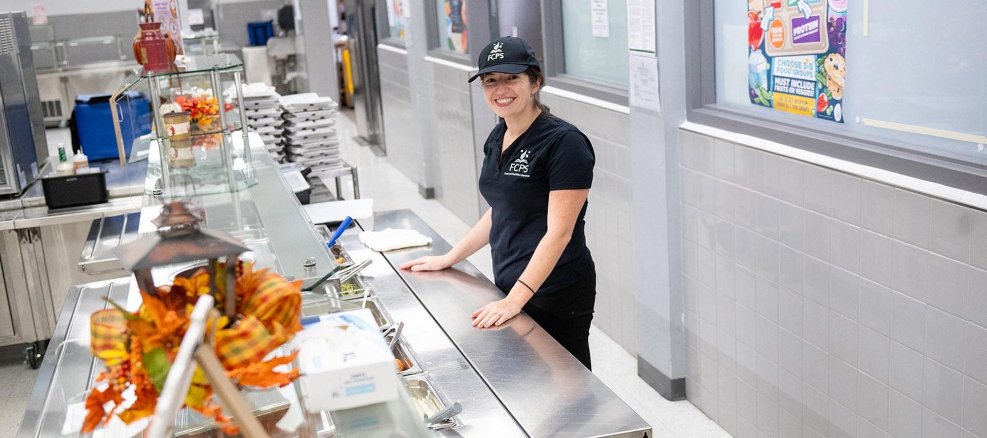 A smiling cafeteria worker in a black uniform stands behind a stainless steel serving counter.