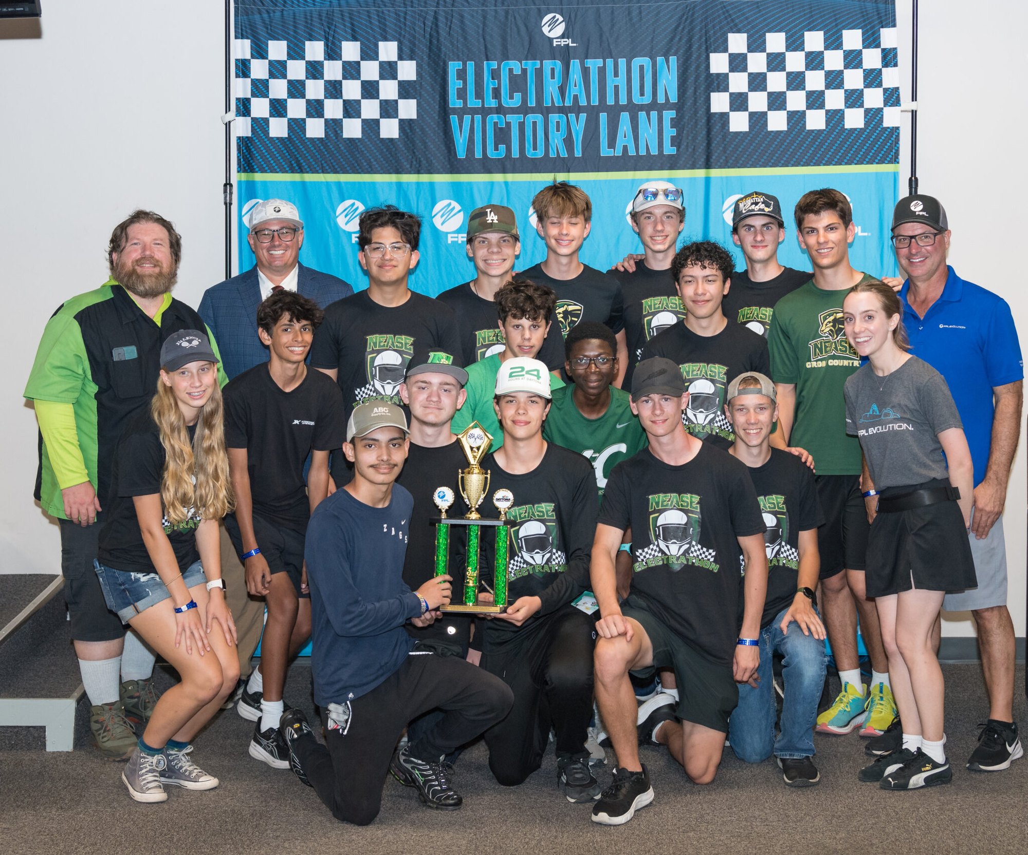 Large group of students holding a trophy in front of a checkered Victory Lane banner.