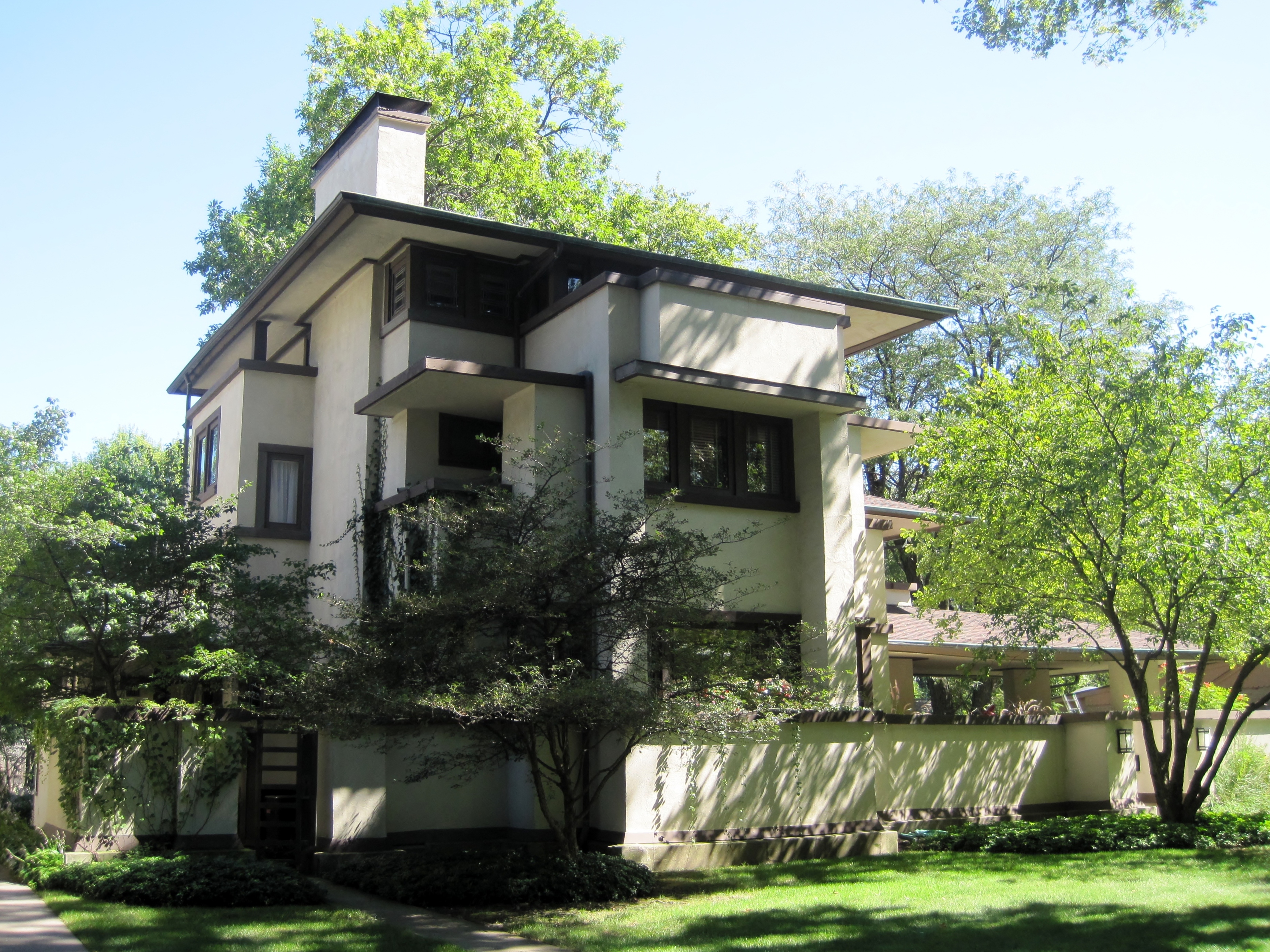 A two-story Prairie Style house with stucco exterior, horizontal banding, and overhanging eaves surrounded by mature trees.