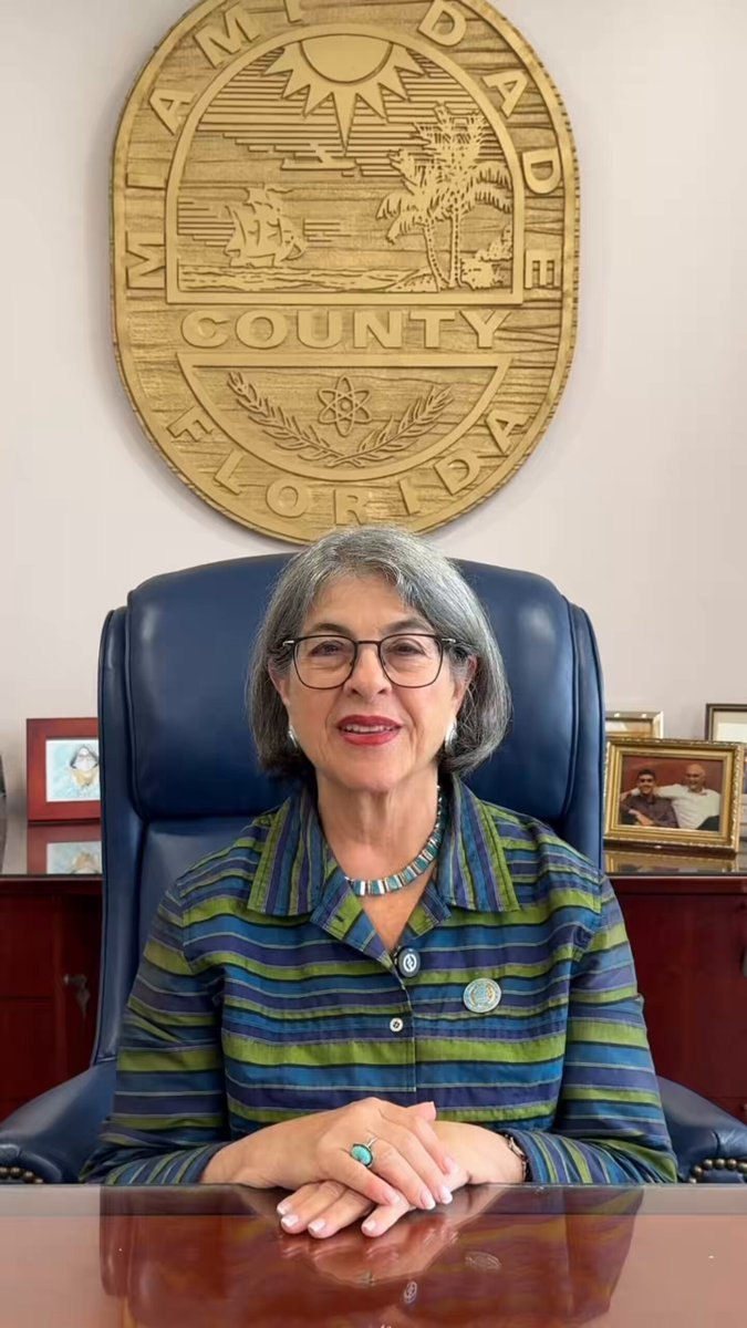 Woman with gray hair and glasses seated at a desk in front of the Miami-Dade County seal.