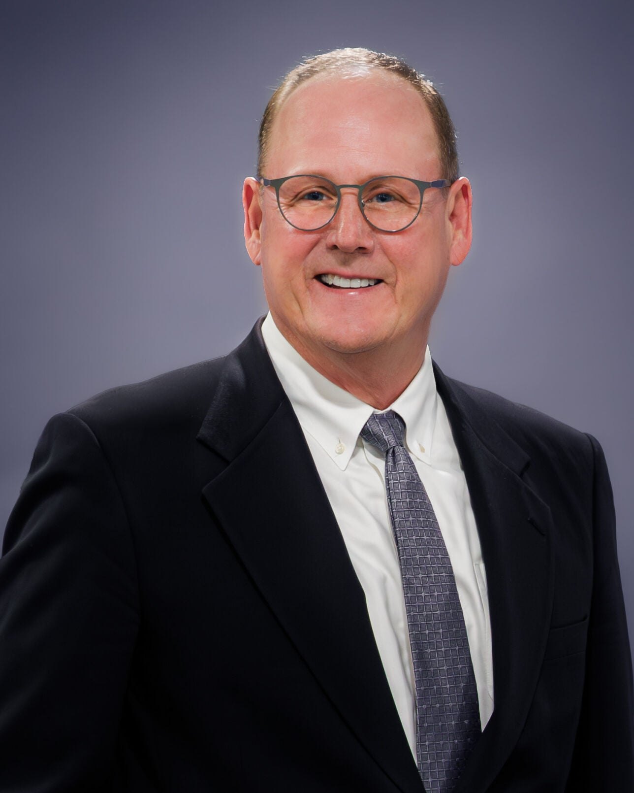 Man with glasses in a dark blazer and gray tie poses for a professional headshot.