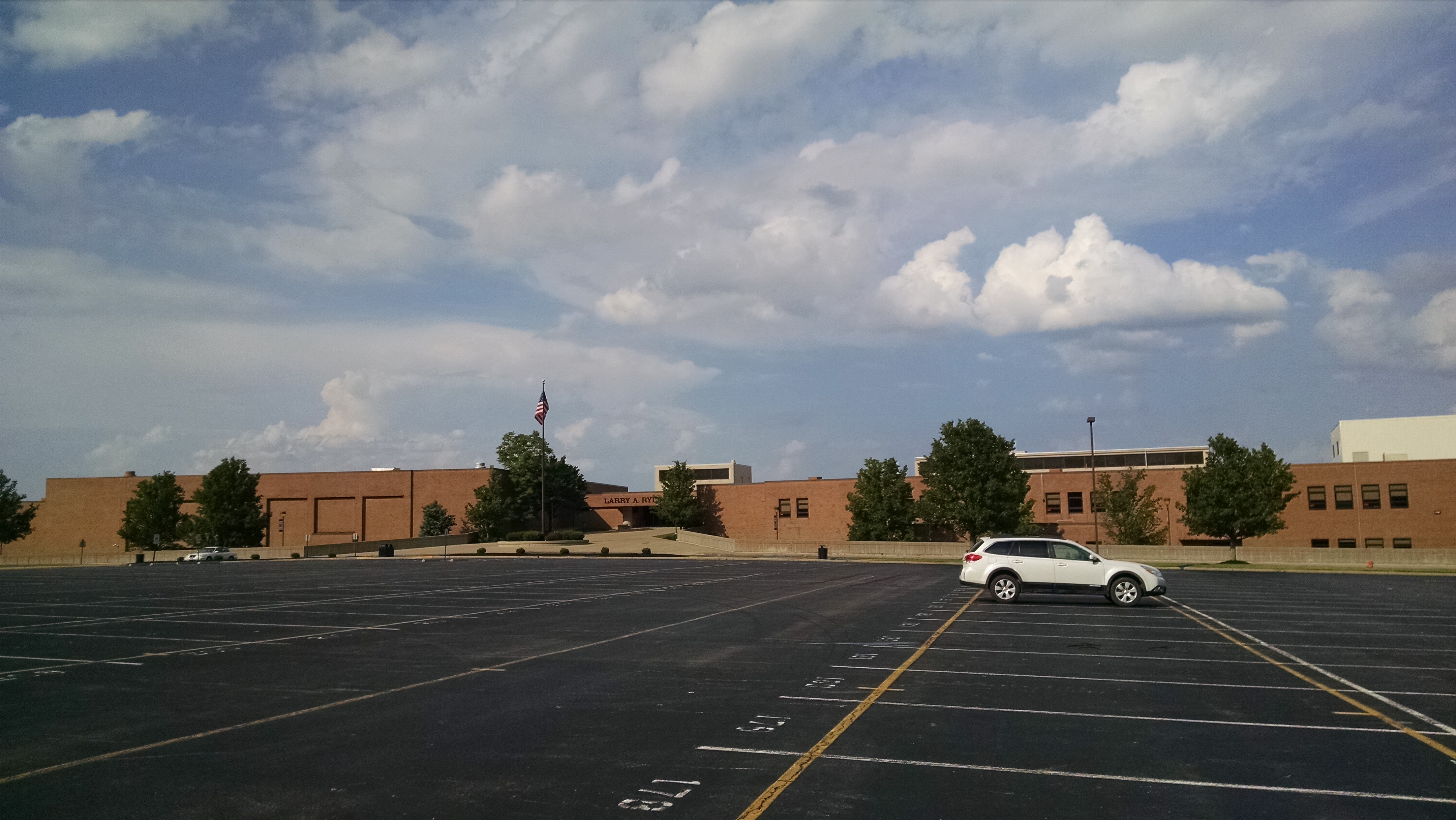 Brick high school building with a flagpole and nearly empty parking lot in front.