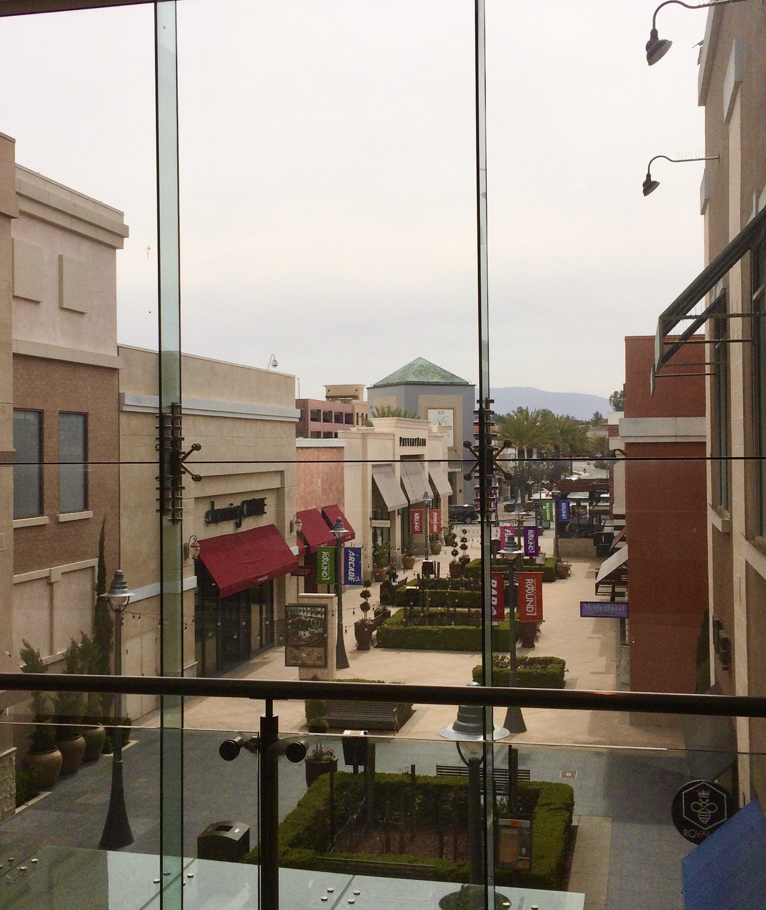 Open-air retail walkway with storefronts, topiary trees, and mountains in the background.
