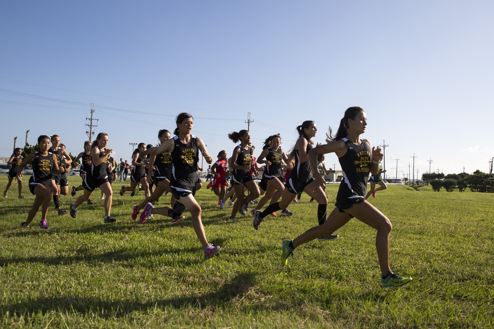 Female high school runners in dark uniforms sprint across a grass field at the start of a cross country race.