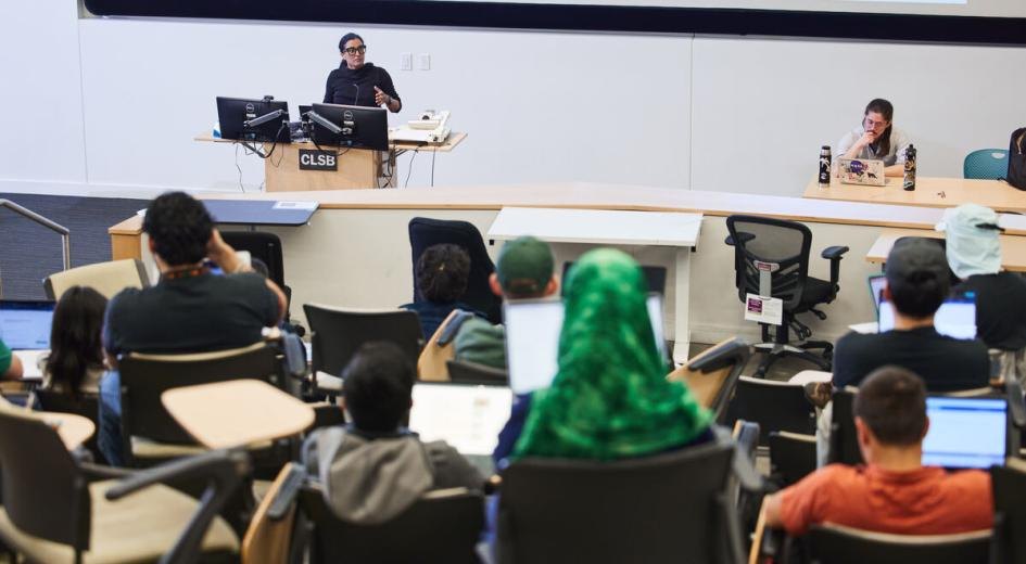 Students with laptops seated in a tiered lecture hall with an instructor at the front.