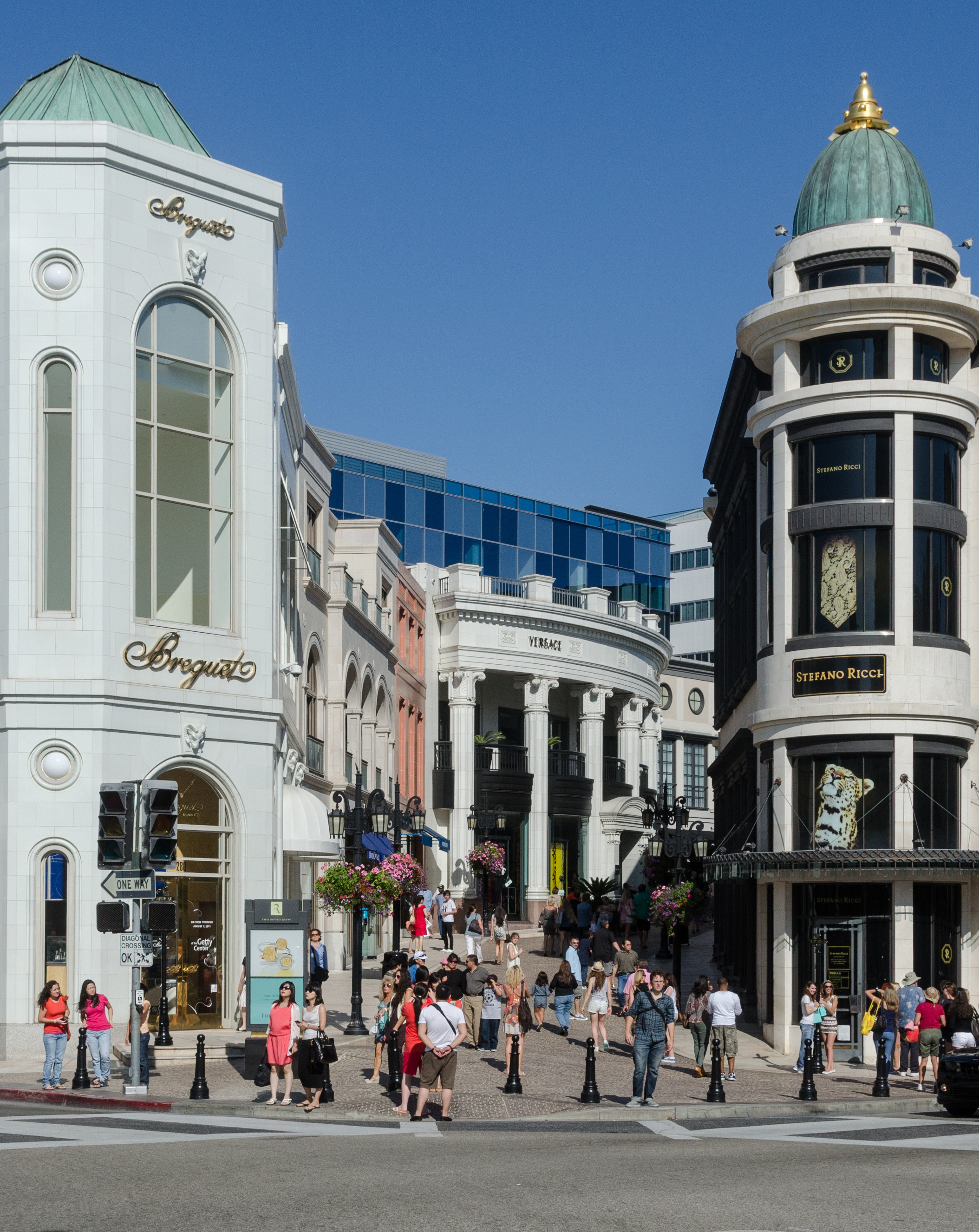 Ornate luxury boutique facades with European-style architecture line a sunny sidewalk.