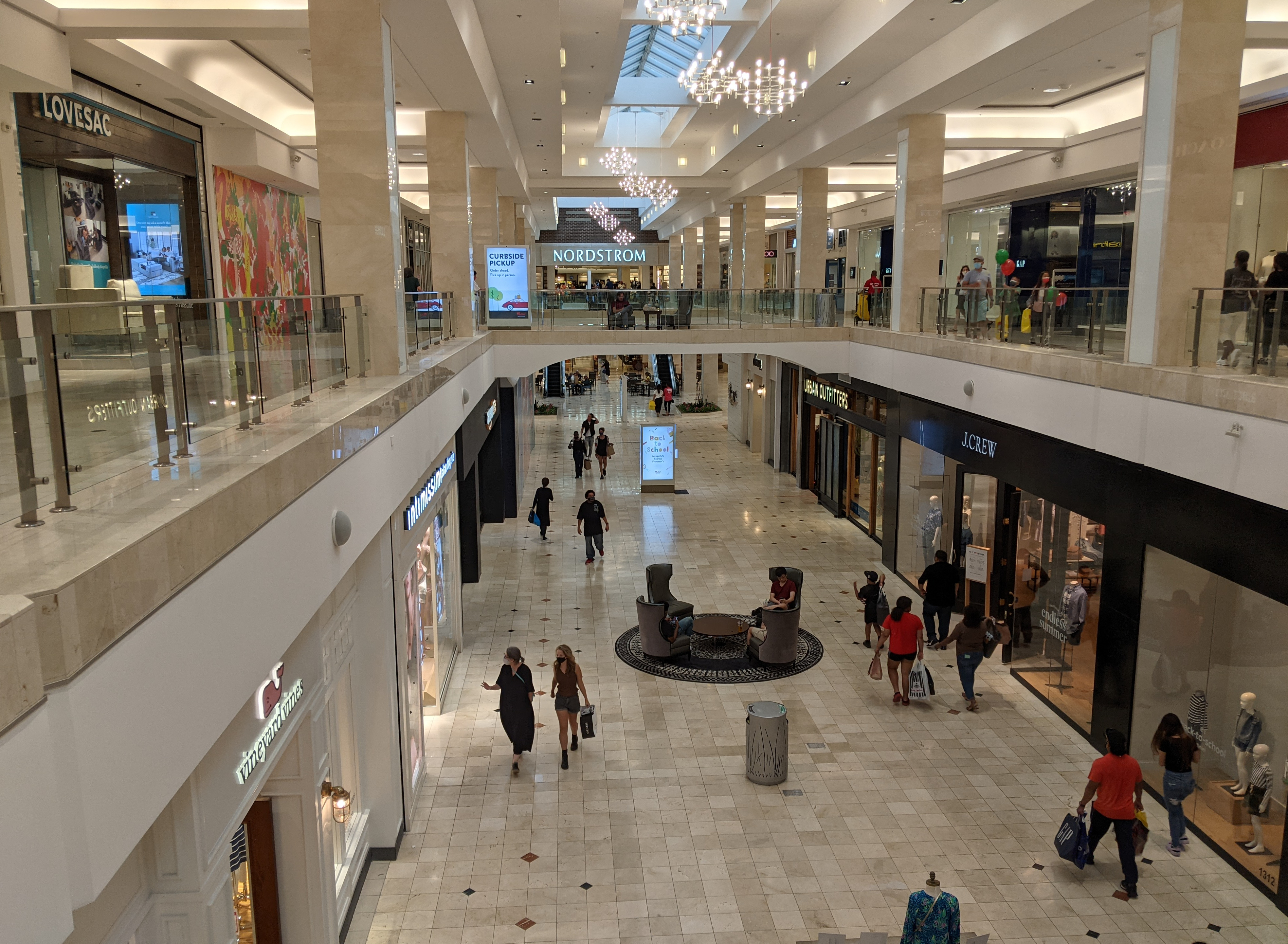 Two-level mall interior with marble floors, white columns, and shoppers walking through a retail corridor.