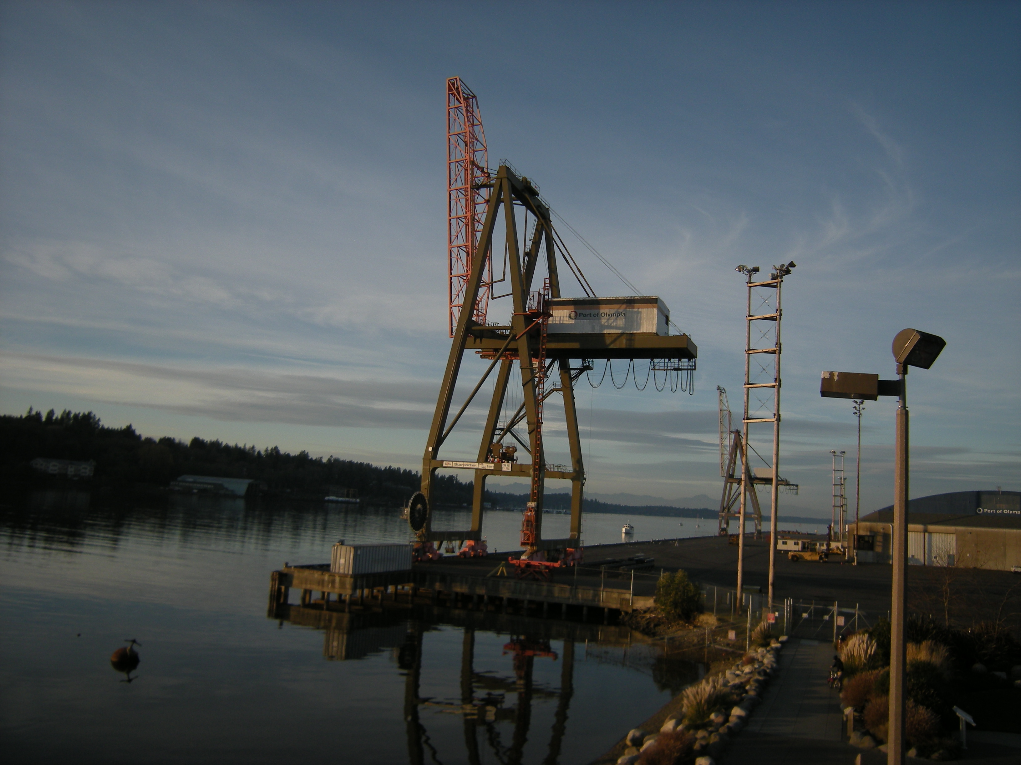 A large industrial crane on a waterfront dock reflected in calm water at dusk.