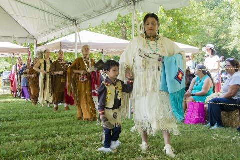 People in traditional Native American regalia walk in a line under a white tent at an outdoor festival.