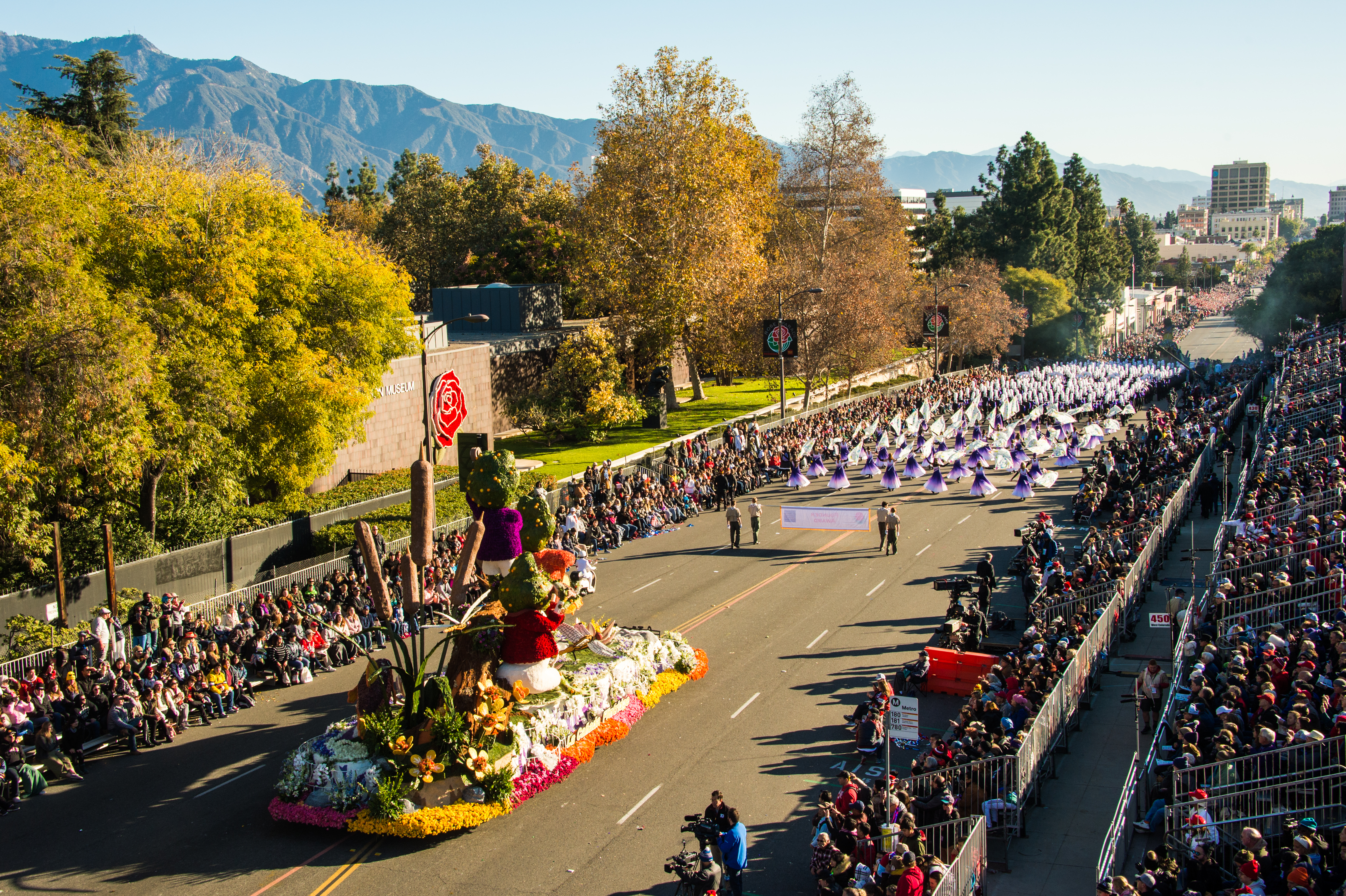 Aerial view of a flower-covered parade float on a crowded boulevard with mountains behind.