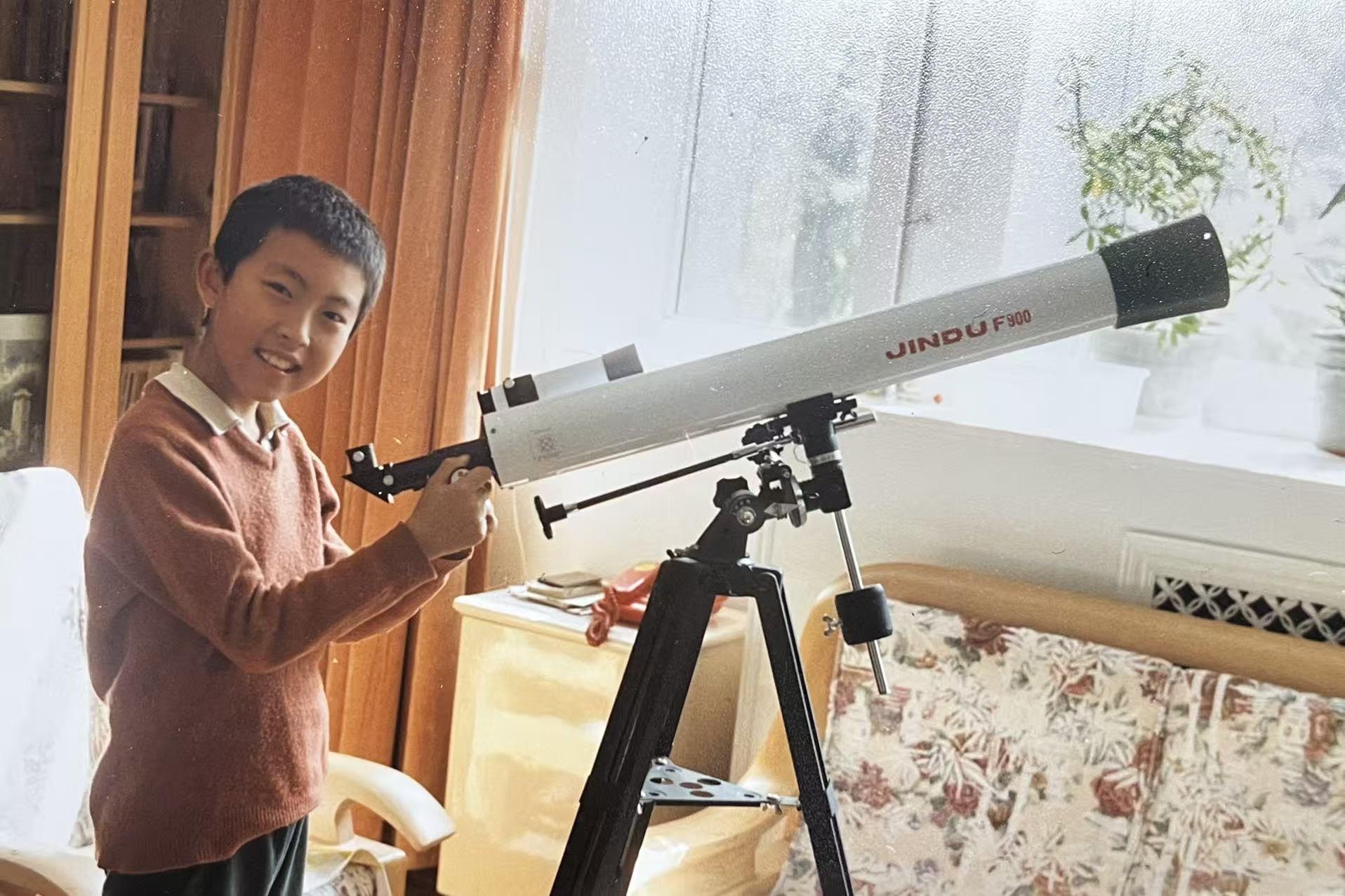 Young boy smiling next to a white refracting telescope on a tripod indoors.