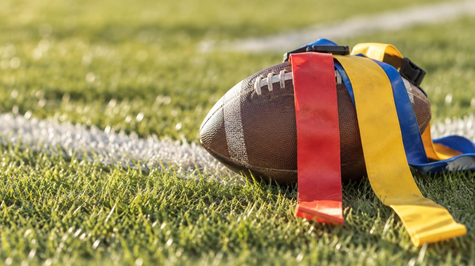 A football with colorful flag belts draped over it resting on green grass near a white yard line.