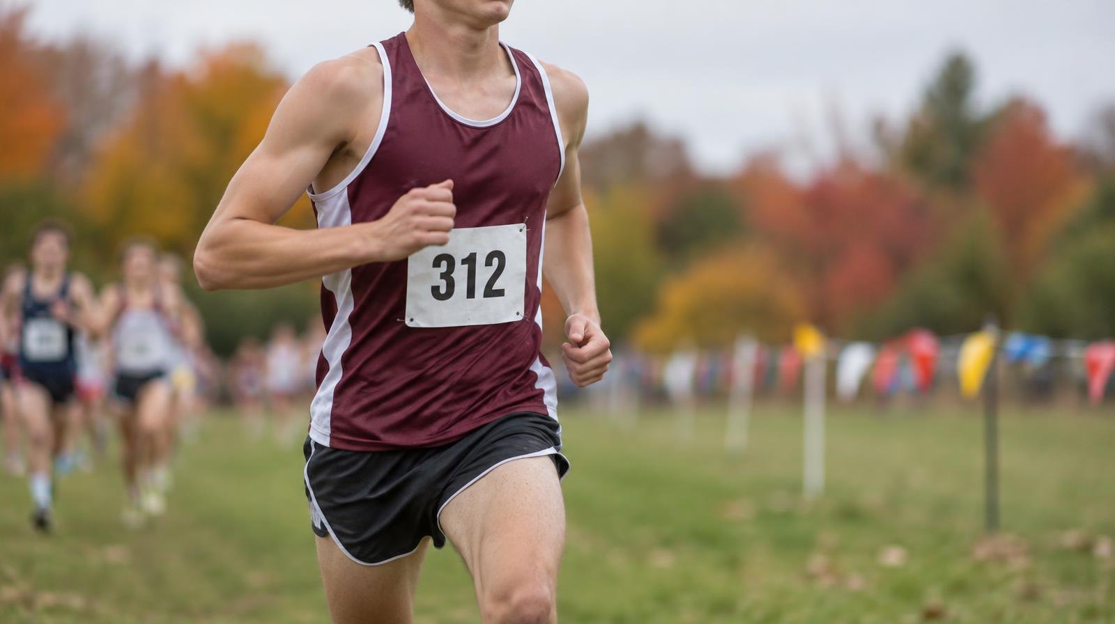A runner in a racing singlet and bib number strides across a grass course with autumn foliage behind.
