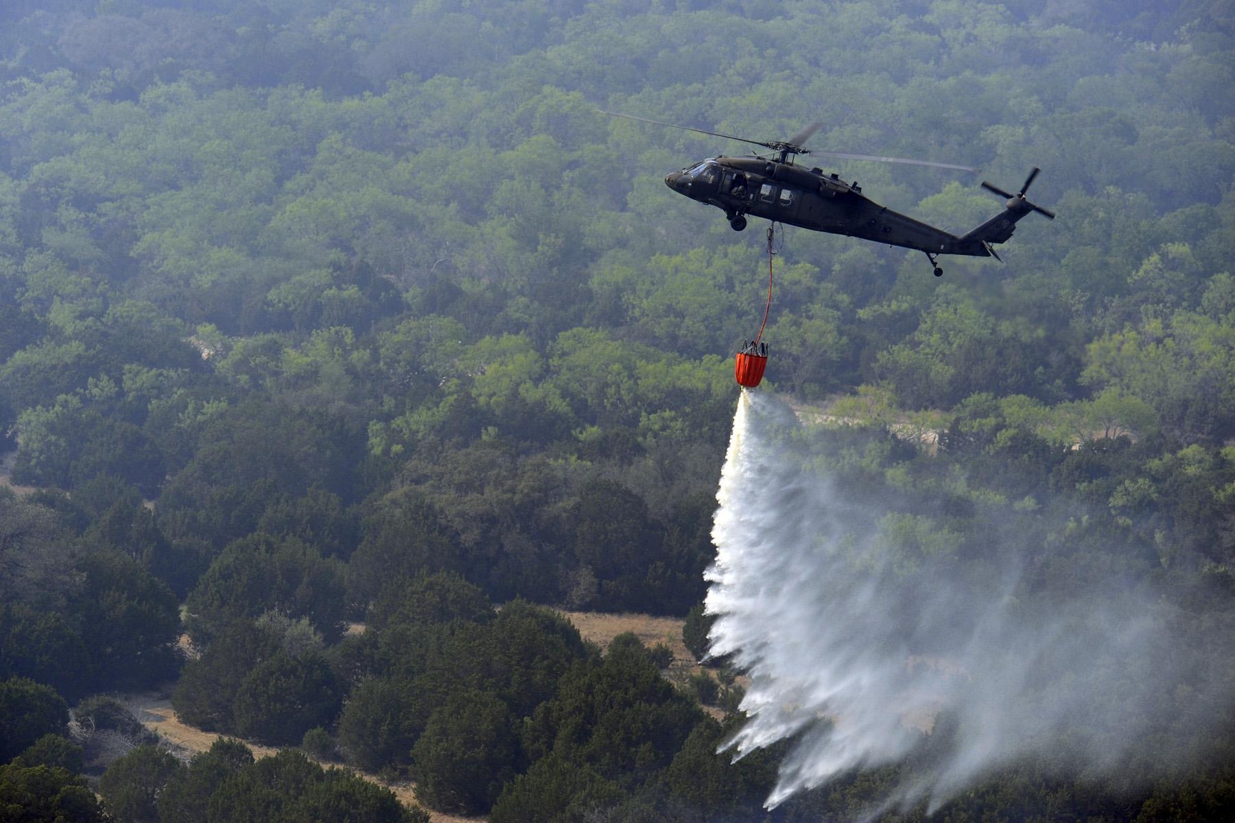 Military helicopter releases water from an orange bucket over a smoky wildfire.