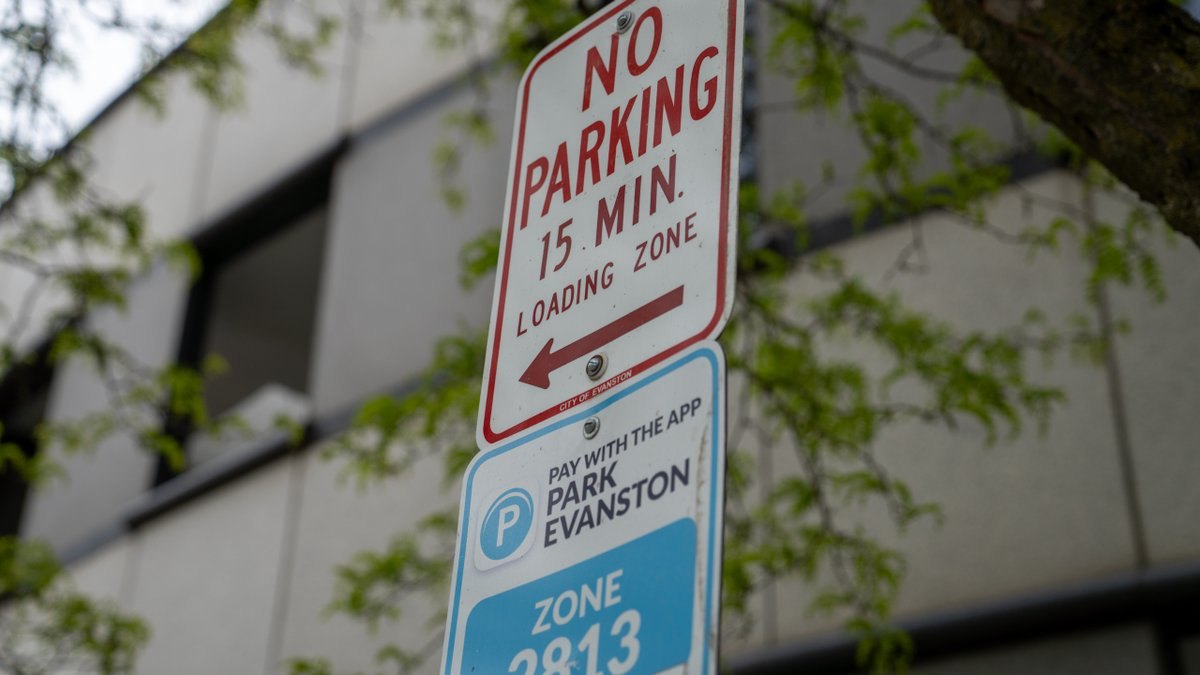 Two parking signs on a pole: a 15-minute loading zone sign above a blue ParkEvanston app payment sign, with trees and a building behind.