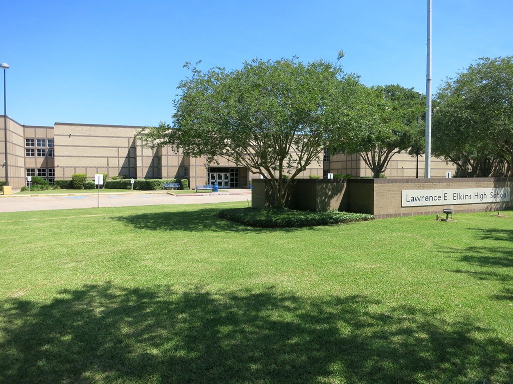 Exterior of a tan brick high school building with a sign, green lawn, and flagpole under a blue sky.