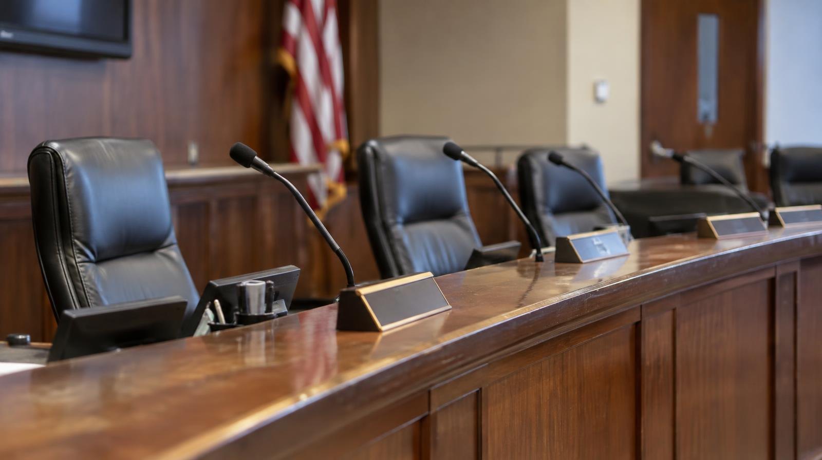 Empty curved council dais with microphones and name placards under overhead lighting.