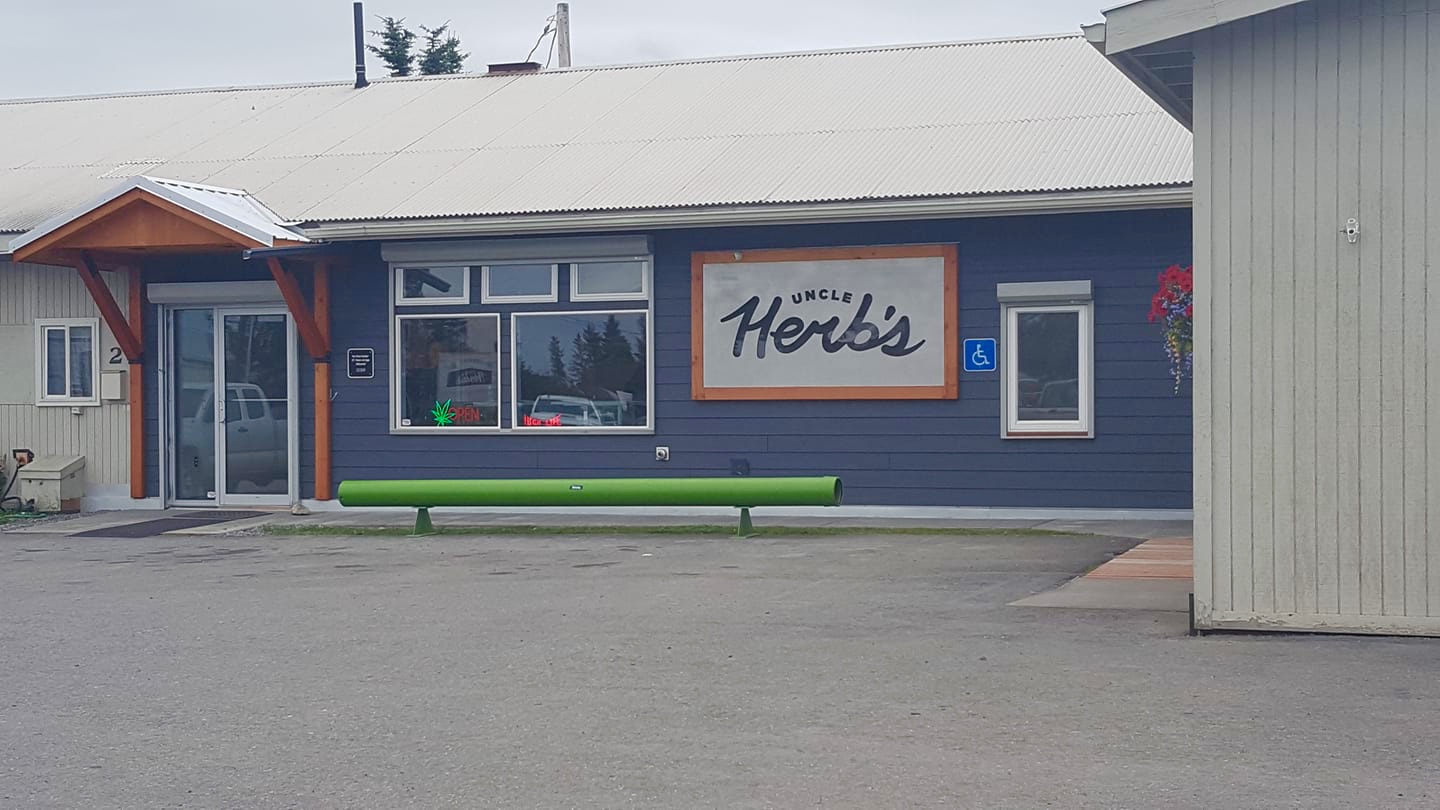 Small cannabis retail store exterior with blue siding and a green bench outside.