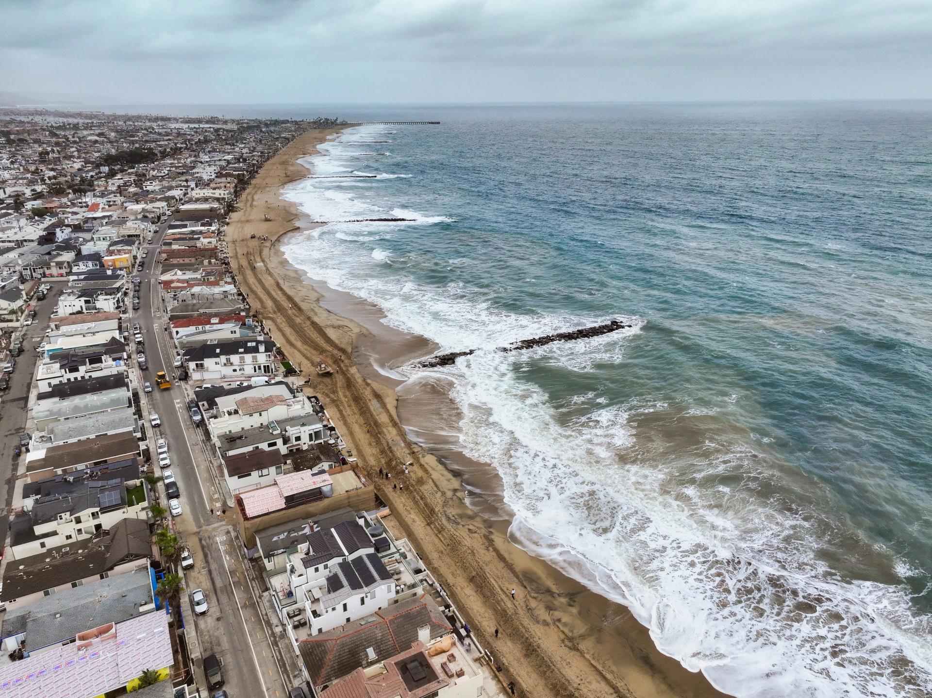 Aerial view of waves breaking along a sandy beach beside rows of homes, with rock groins extending into the ocean under an overcast sky.