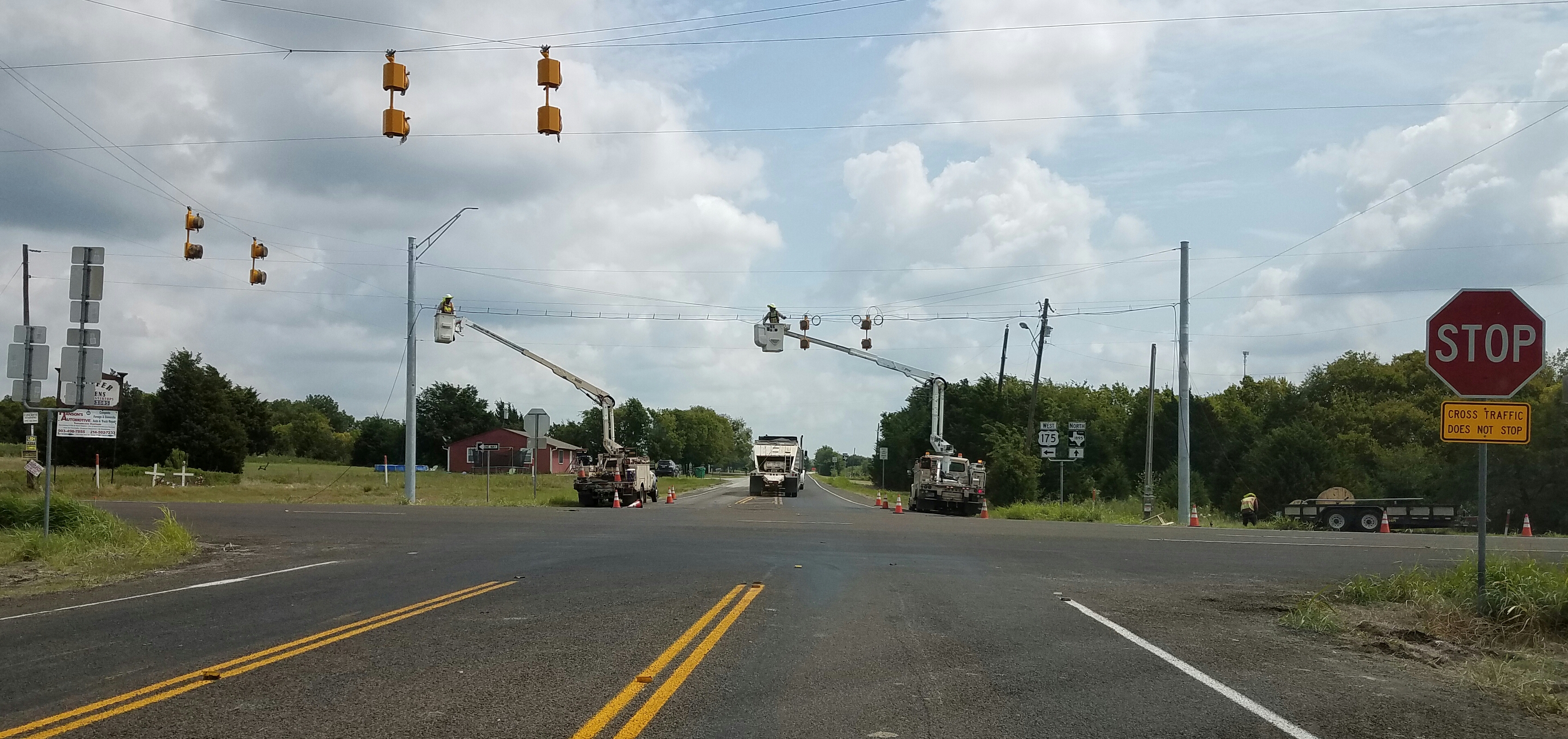 Two bucket trucks with workers installing traffic signal heads at a rural road intersection.