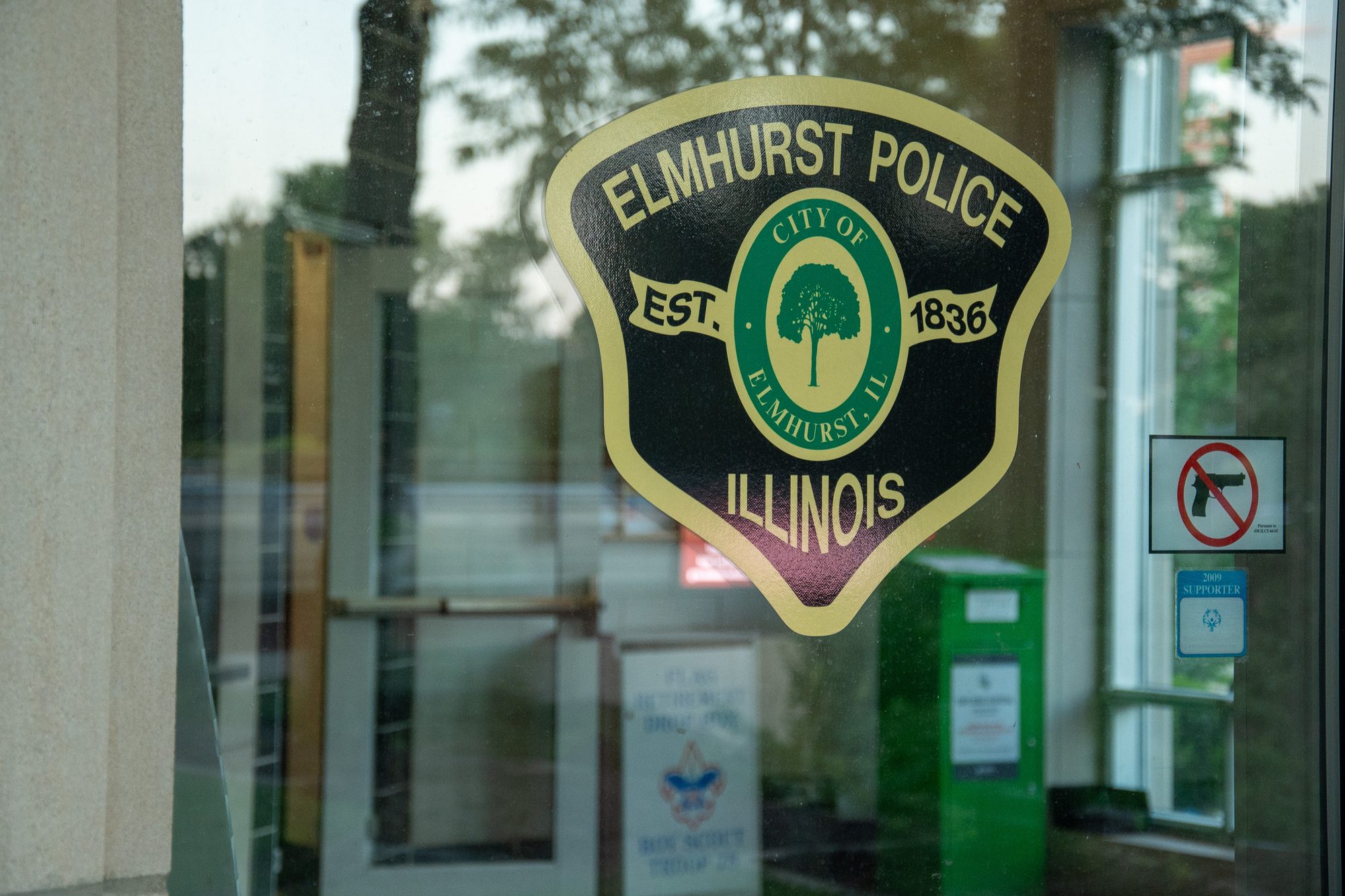 Elmhurst police station. A close-up of the Elmhurst Police Department badge decal affixed to a glass door or window, showing the shield-shaped logo with 'ELMHURST POLICE ILLINOIS' and 'EST. 1836' text surrounding a City of Elmhurst seal. A no-guns sign is visible to the right on the glass.