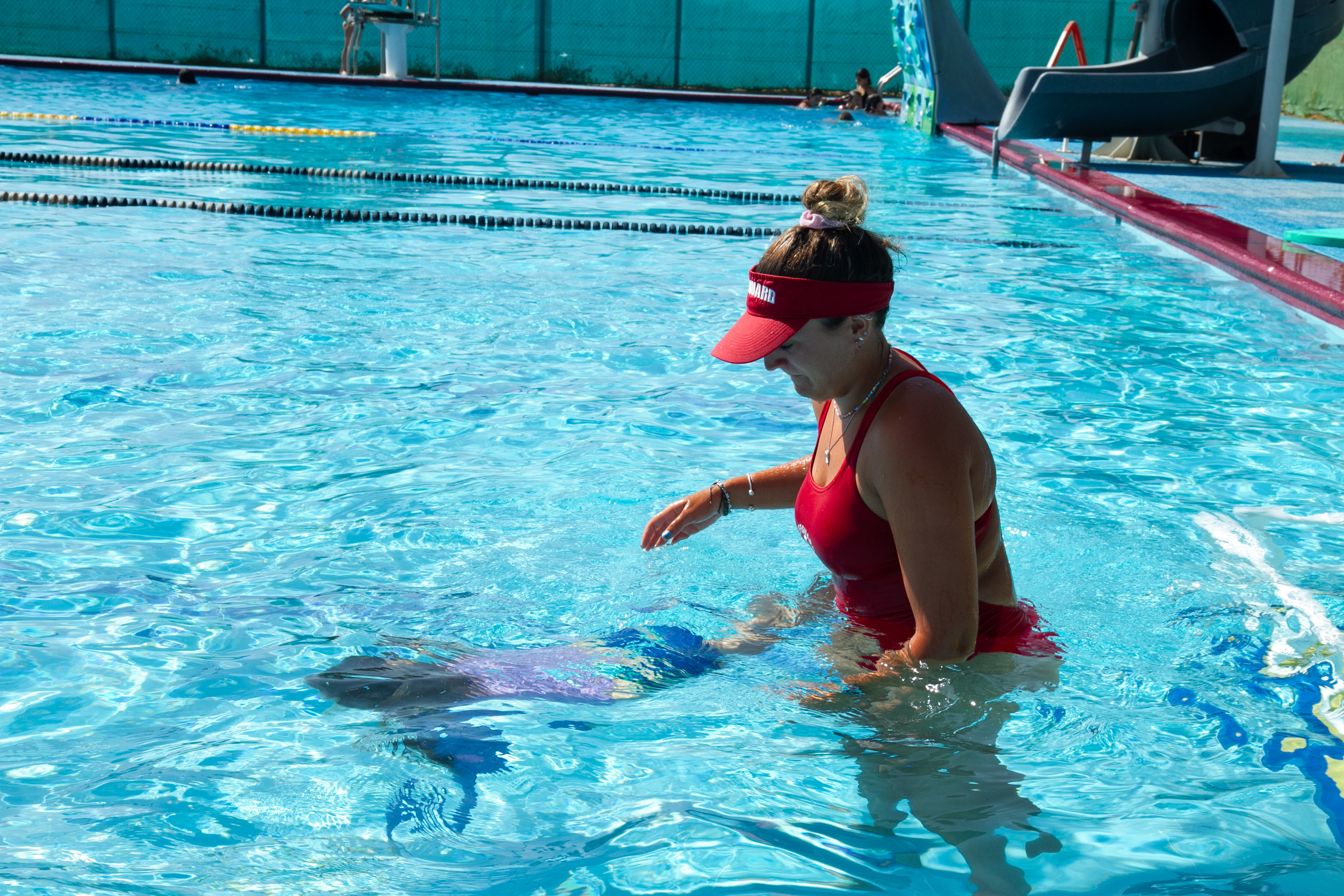 A swim instructor in a red swimsuit stands in a pool watching a child swim beside her.