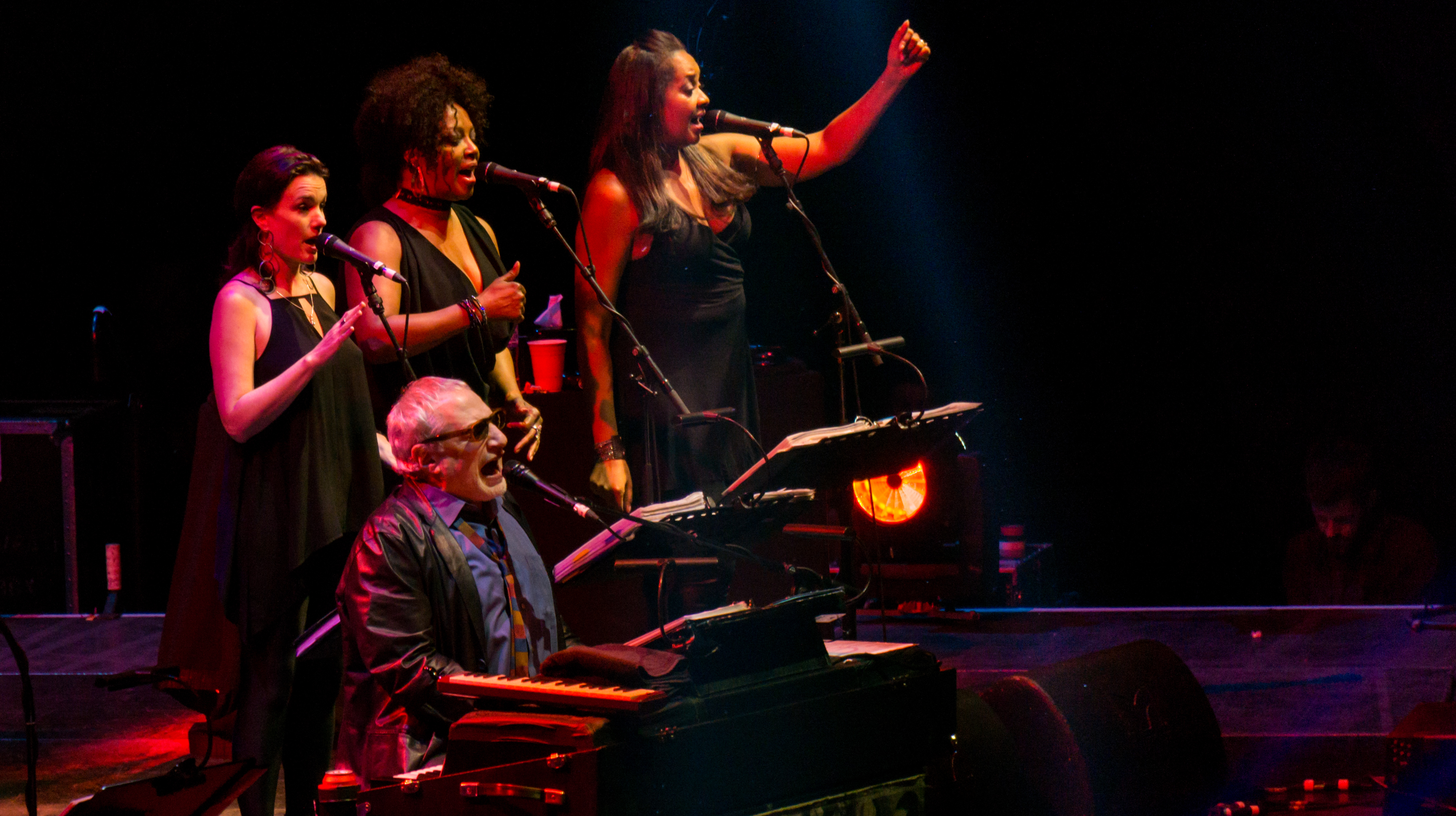 A keyboardist and three vocalists perform on a dimly lit concert stage.