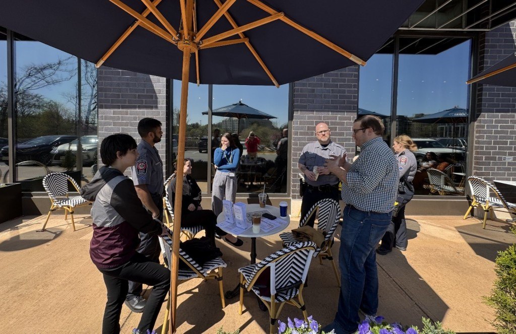 People standing and talking at an outdoor patio under a large umbrella with bistro tables.