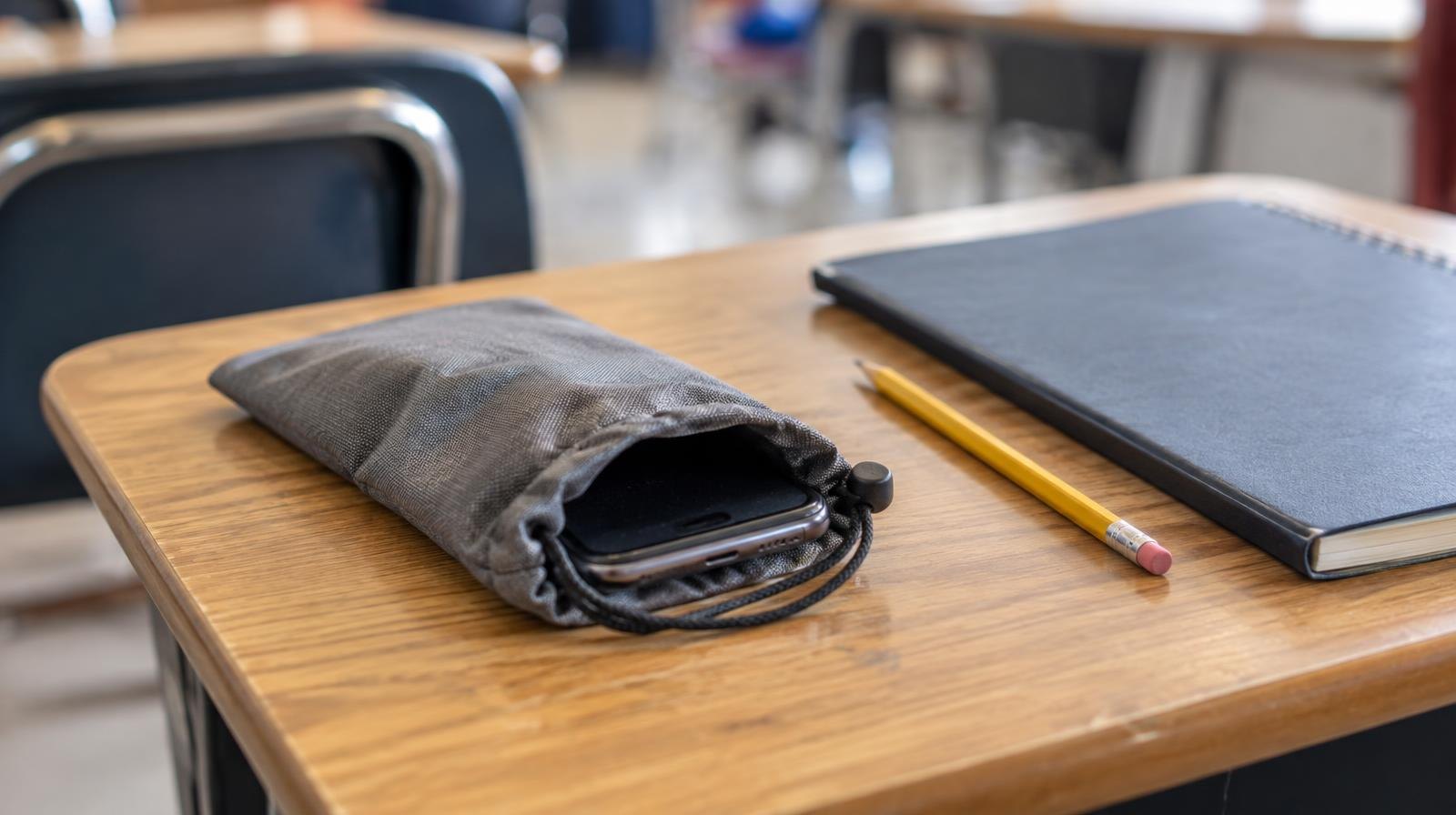 A smartphone in a locked fabric pouch on a desk beside a notebook and pencil.
