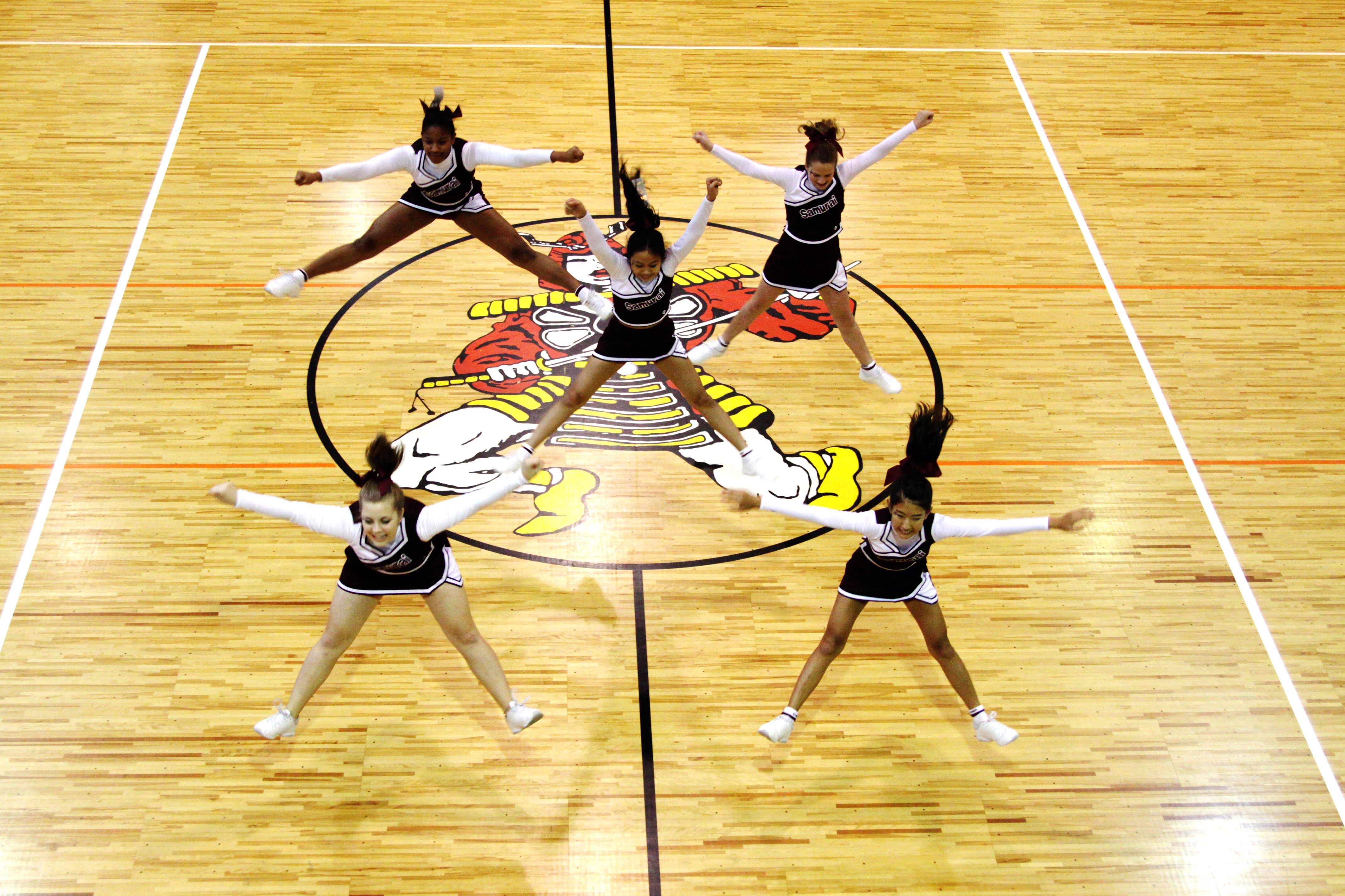 Seven cheerleaders in uniforms performing synchronized jumps on a gymnasium floor.