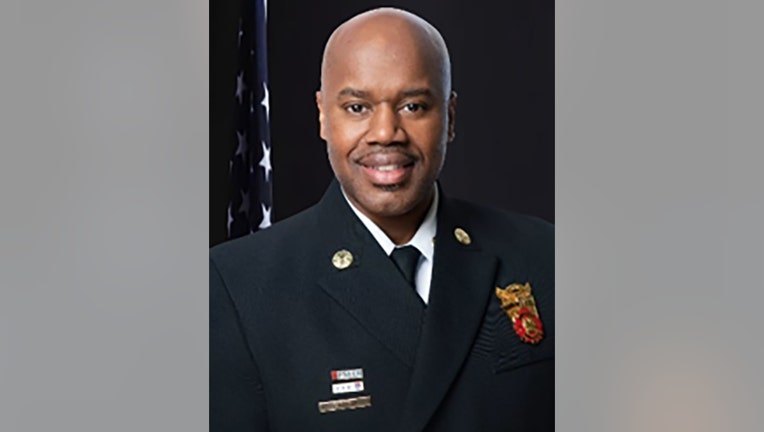 Man in dark fire department dress uniform with badge and rank insignia, posed formally.