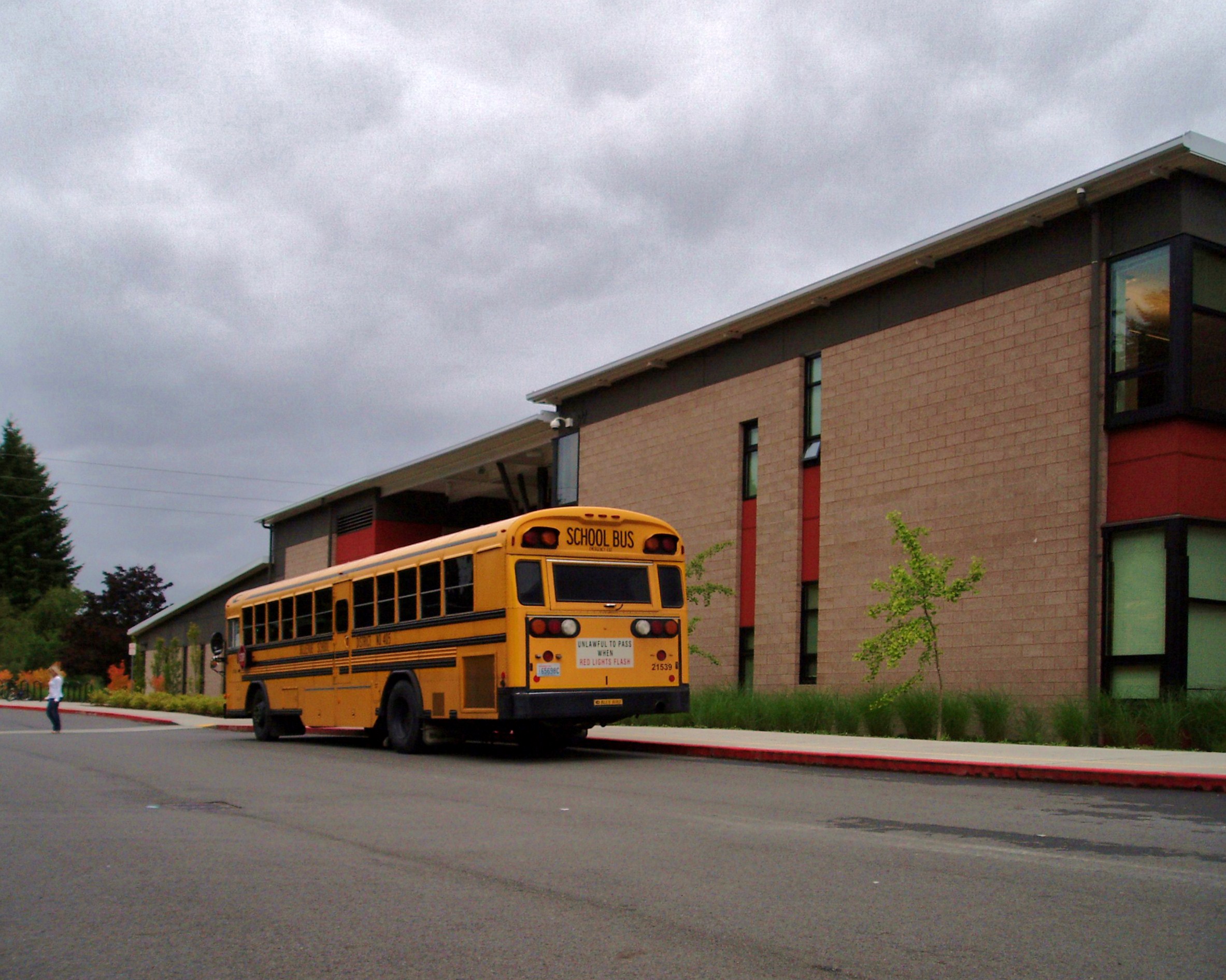 Yellow school bus parked in front of a brick school building under overcast skies.