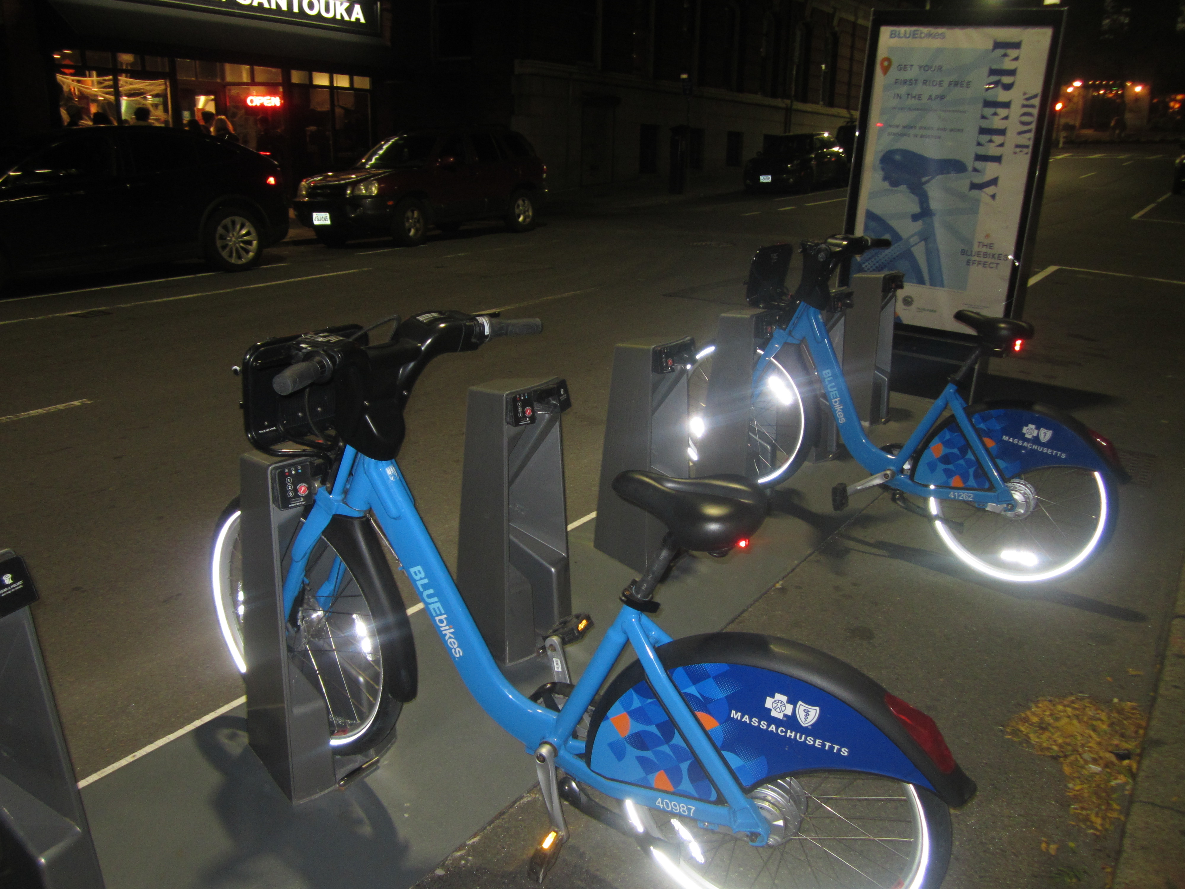 Blue shared bicycles docked at a bike-share station at night.
