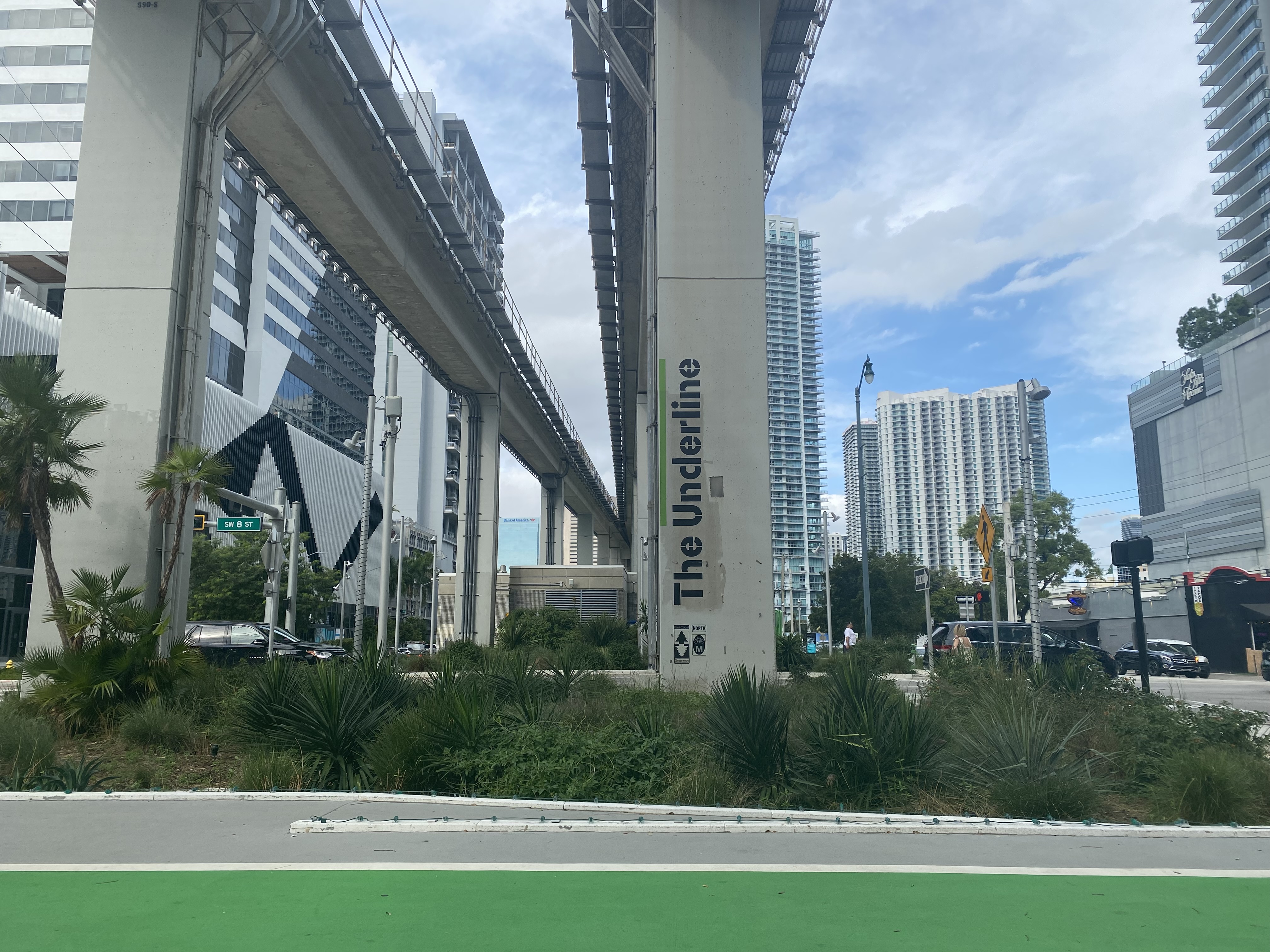 Paved trail beneath an elevated rail structure with a column labeled 'The Underline' and palm trees.